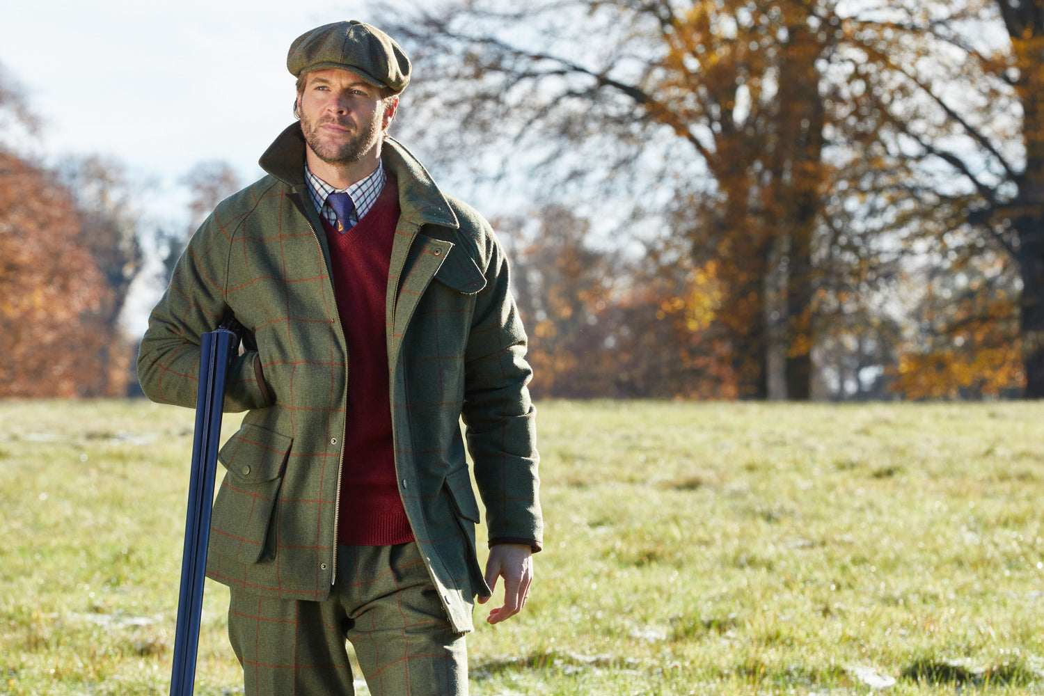 Man in a tweed jacket and flat cap standing in an open field with trees in the background.