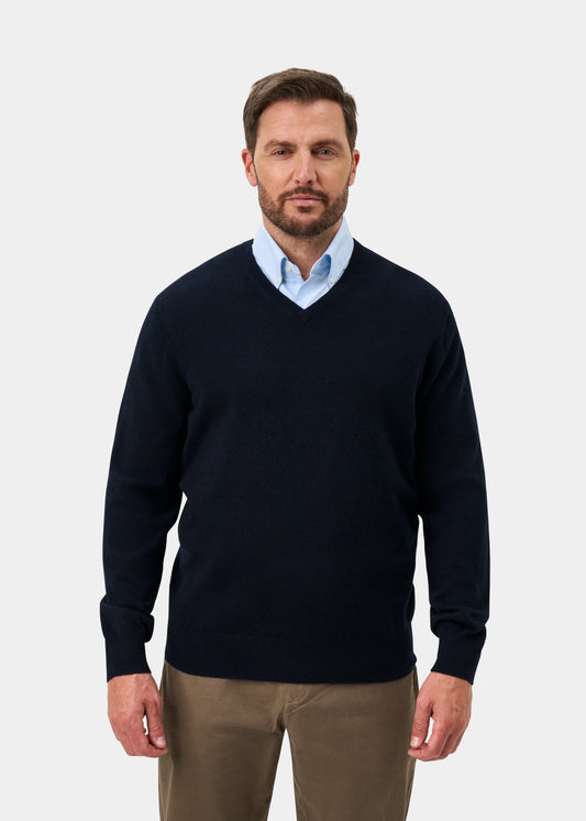 Man in Haddington Pure Cashmere V-Neck Jumper in Dark Navy, light blue shirt, khaki pants; plain background.