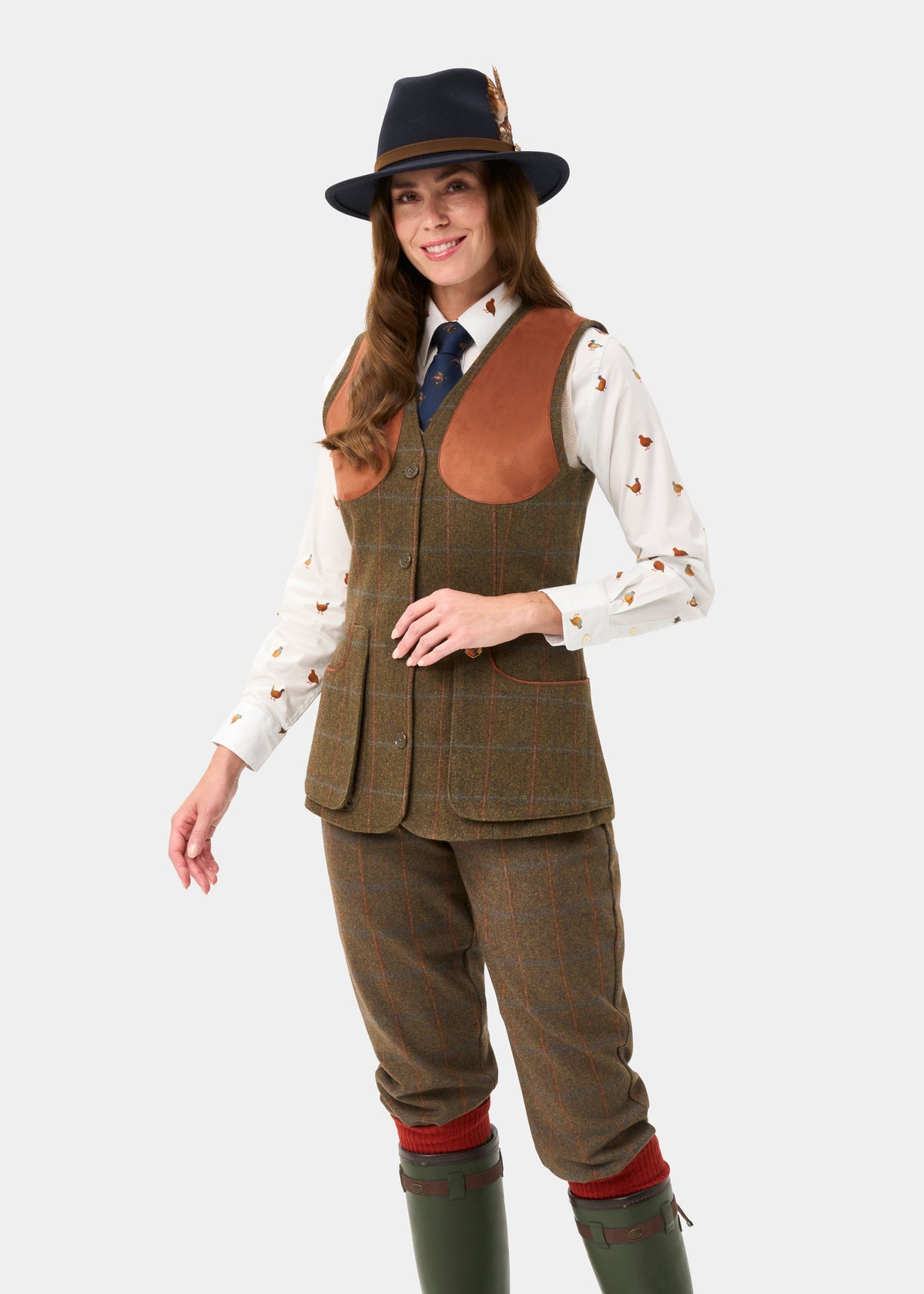 Woman in Combrook Ladies Tweed Water Repellent Waistcoat (Willow), hat, tie & green boots, smiling against plain backdrop.