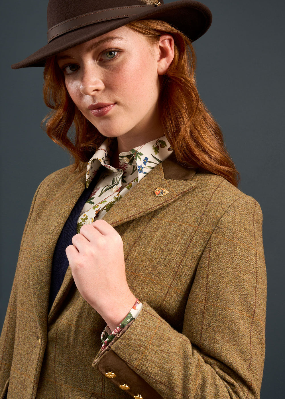 Woman wears Combrook Ladies Tweed Water Repellent Coat in Grove, floral shirt peeking out, against dark backdrop.