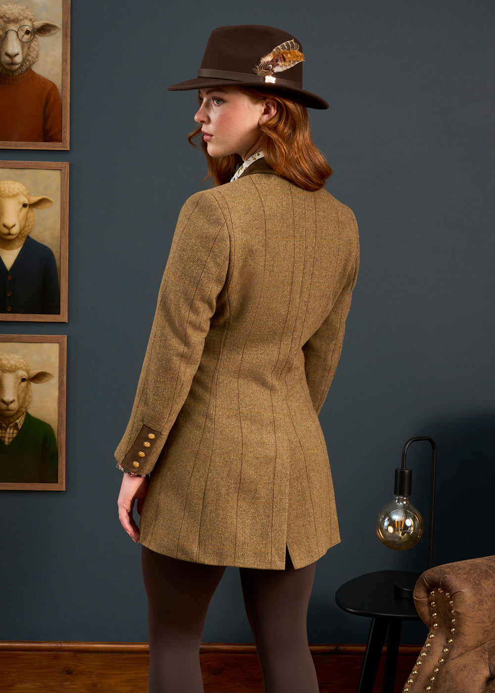 Woman in brown feathered hat and Combrook Ladies Tweed Coat poses in vintage room with sheep portraits.