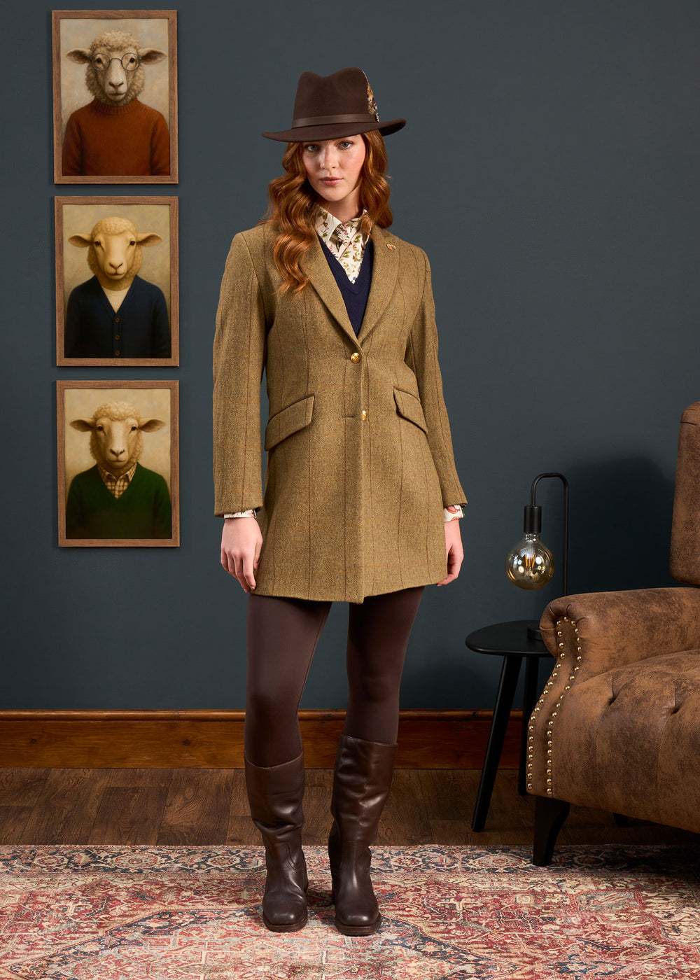 A woman in a Combrook Ladies Tweed Coat stands indoors; behind her are three sheep portraits dressed like people.