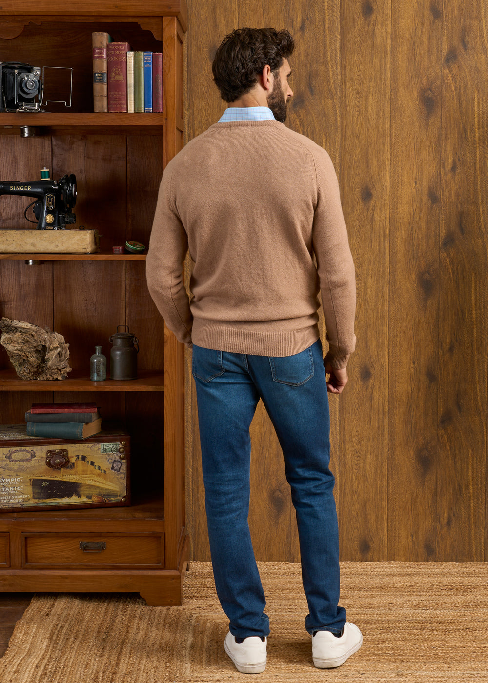 A man in a Hampshire Lambswool Vee Neck Jumper in Camel and blue jeans faces a wooden bookshelf with vintage items.