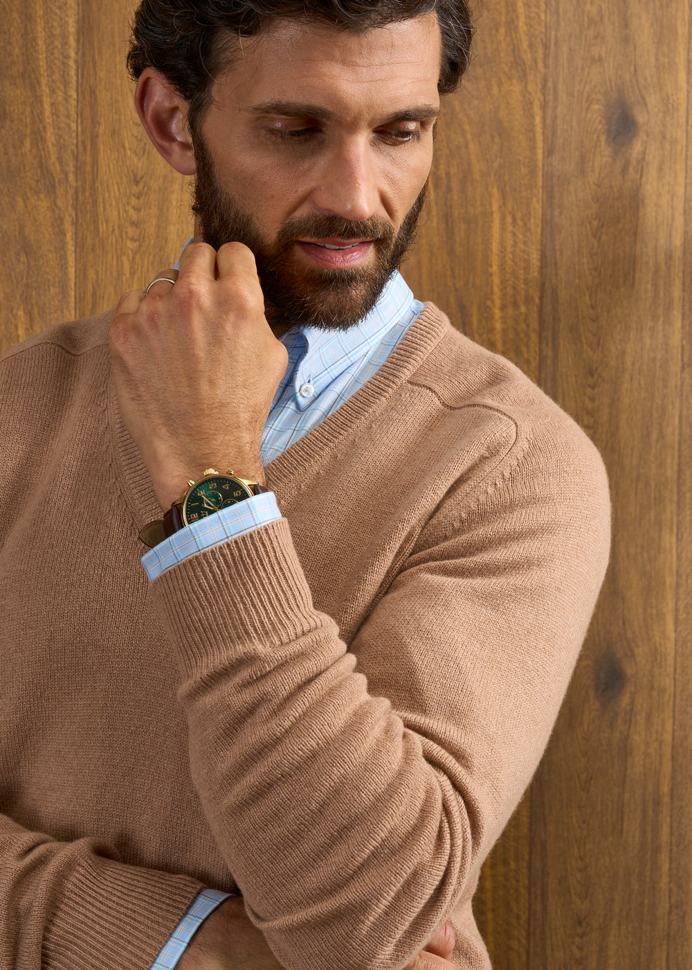 Man wears Hampshire Lambswool Vee Neck Jumper in Camel over blue shirt, gold watch visible, wood panel backdrop.