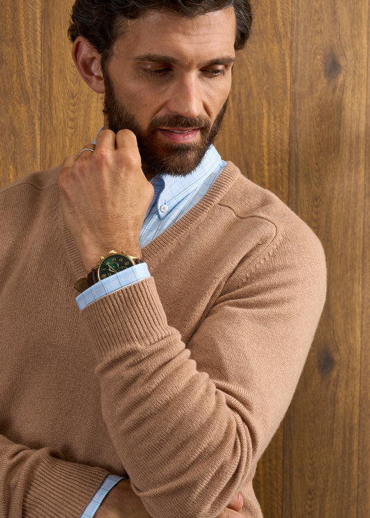 Man wears Hampshire Lambswool Vee Neck Jumper in Camel over blue shirt, gold watch visible, wood panel backdrop.