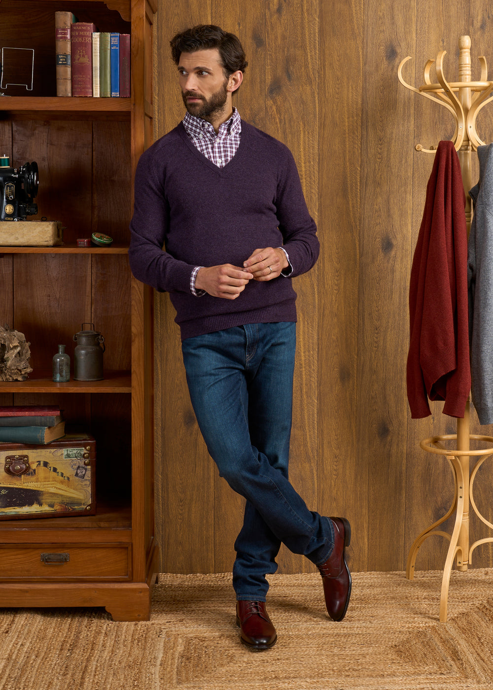 Man in elderberry Hampshire lambswool V-neck jumper and jeans stands by wooden shelf and coat rack with red sweater.