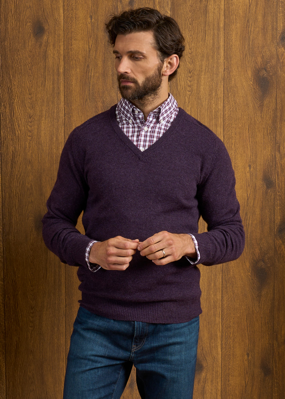 Man in a Hampshire Elderberry Lambswool Vee Neck Jumper stands by a wooden wall, looking to his left.