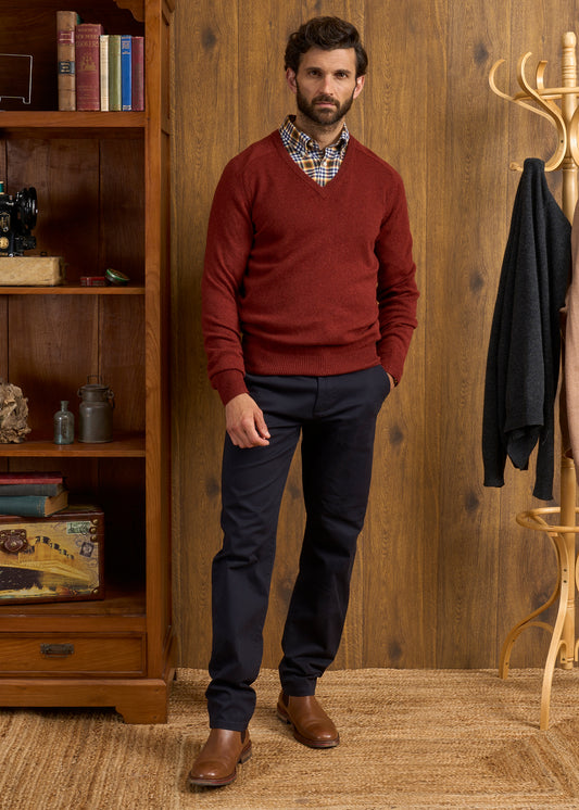 Man in Hampshire Lambswool Vee Neck Jumper in Sienna, plaid shirt, navy pants stands by rack and bookshelf indoors.