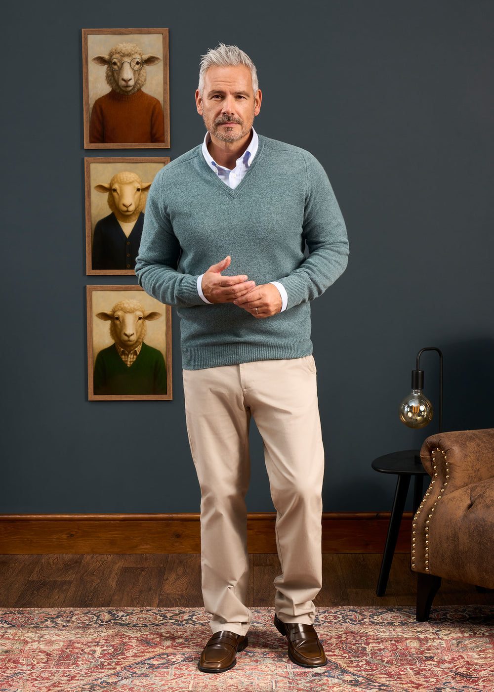 A man wears a Hampshire Lambswool Vee Neck Jumper in Caspian, standing by a chair and sheep portraits indoors.