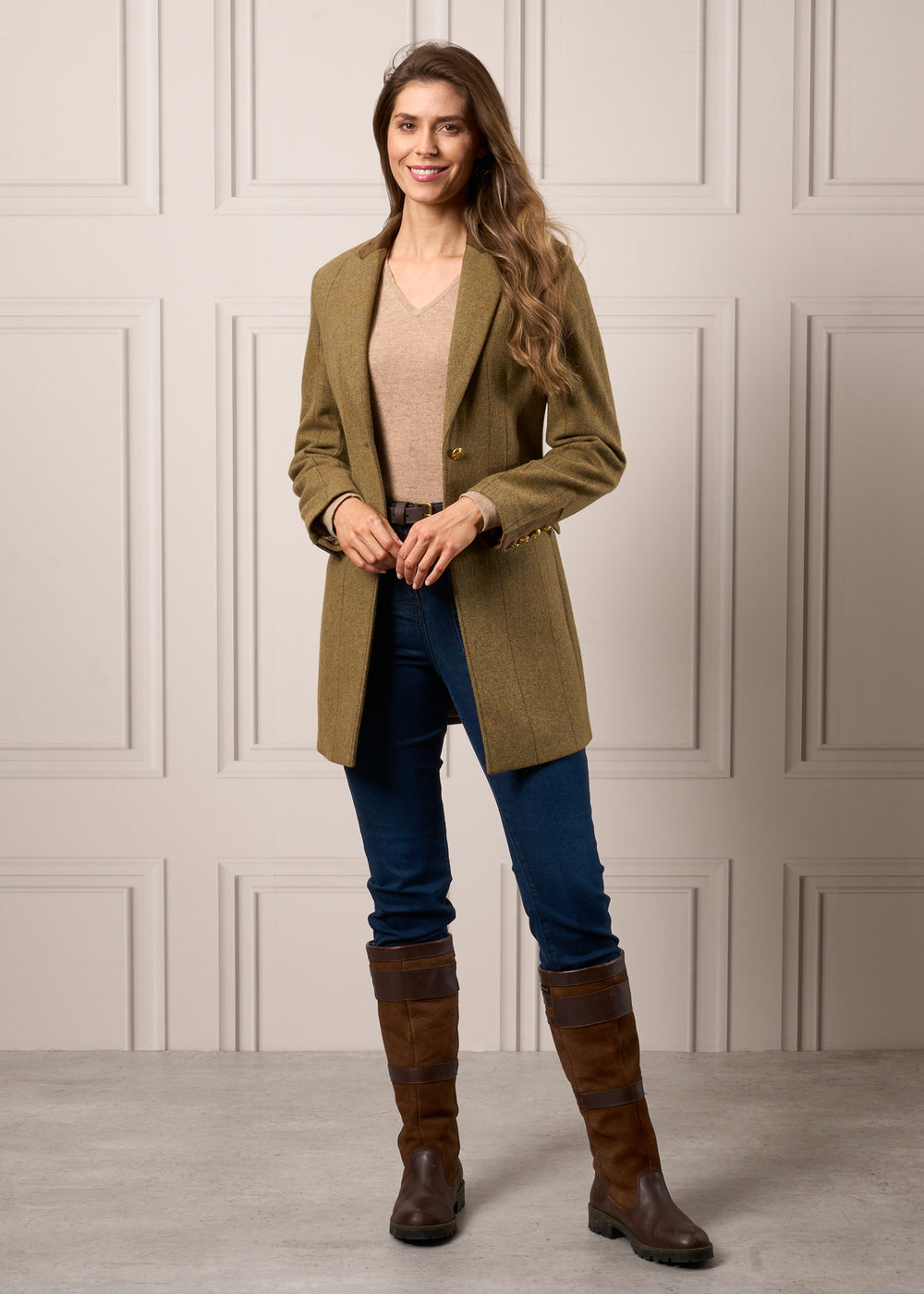 Woman in Combrook Ladies Tweed Mid-Thigh Coat (Grove), beige sweater, blue jeans, and boots before a beige paneled wall.