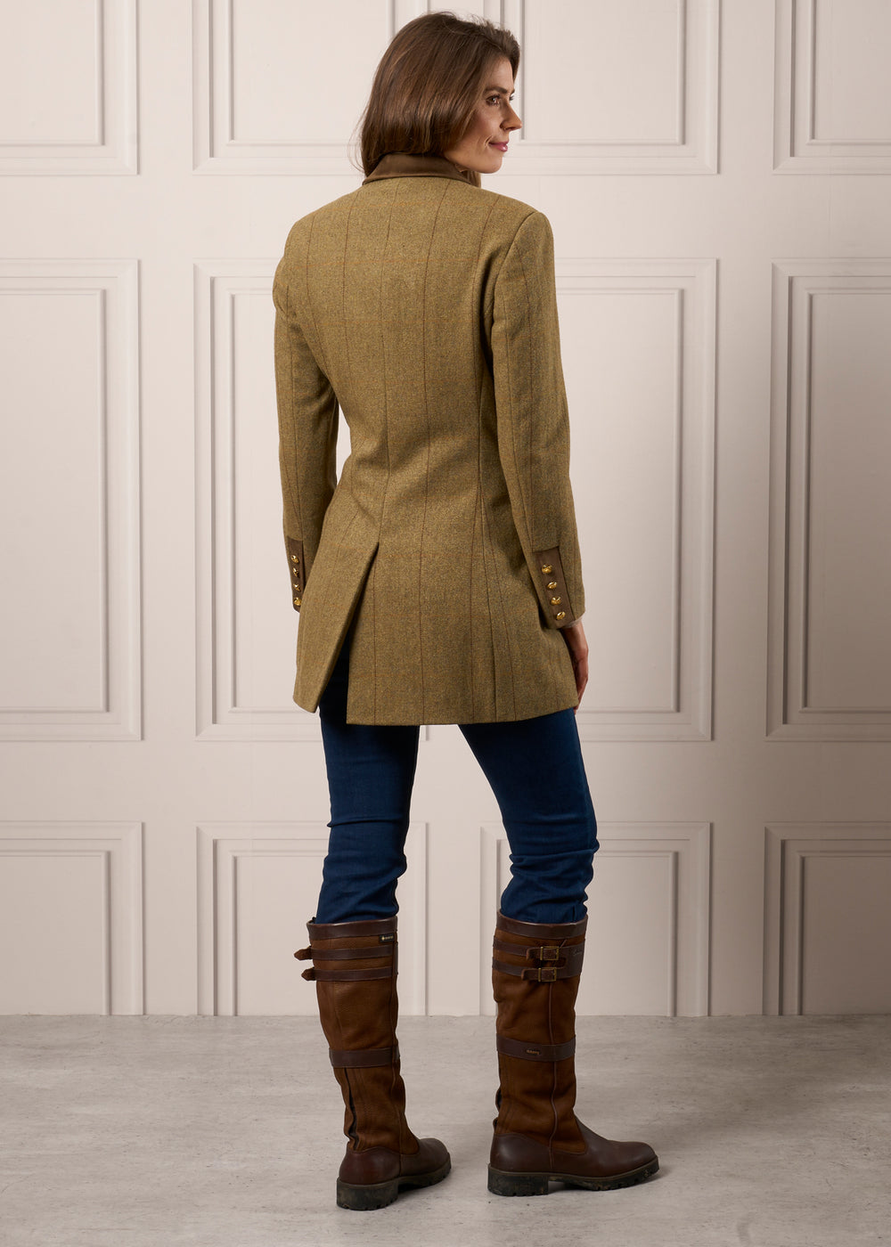 Woman in Combrook Ladies Tweed Mid-Thigh Coat (Grove), blue jeans, and brown riding boots stands in a paneled room.
