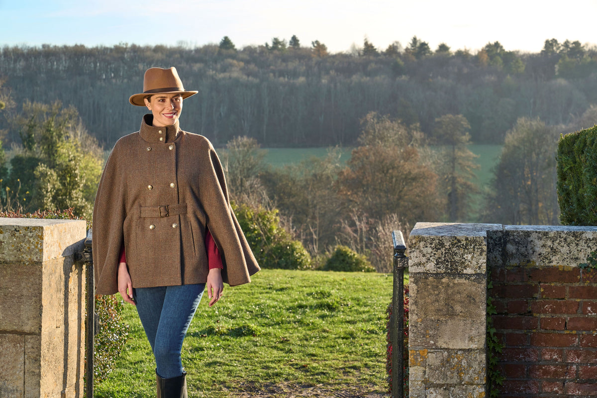 A woman wearing a brown wool cape jacket, brown hat, blue jeans, and black boots stands in a stone gate on a grassy hill with a forested landscape in the background.