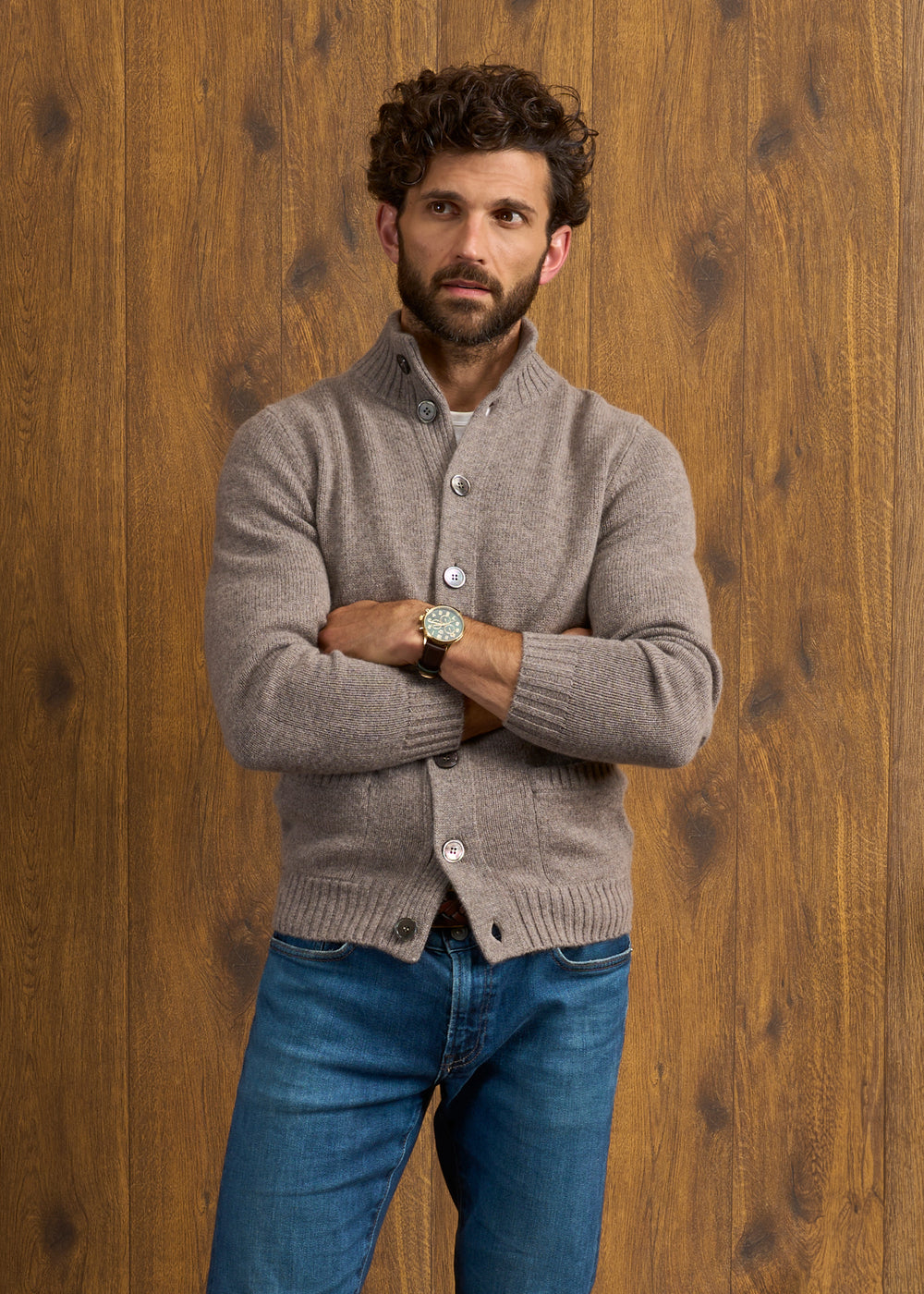 Man with curly hair in a Vole Landford Lambswool Mock Neck Jumper stands arms crossed before a wooden wall.