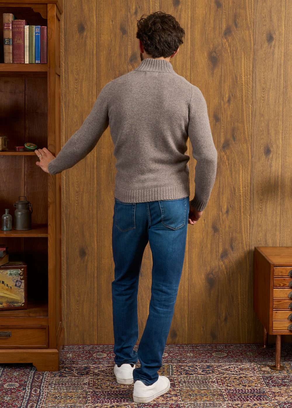 Man in Landford Lambswool Buttoned Mock Neck Jumper (Vole) reaches toward a wooden bookcase with his left hand.