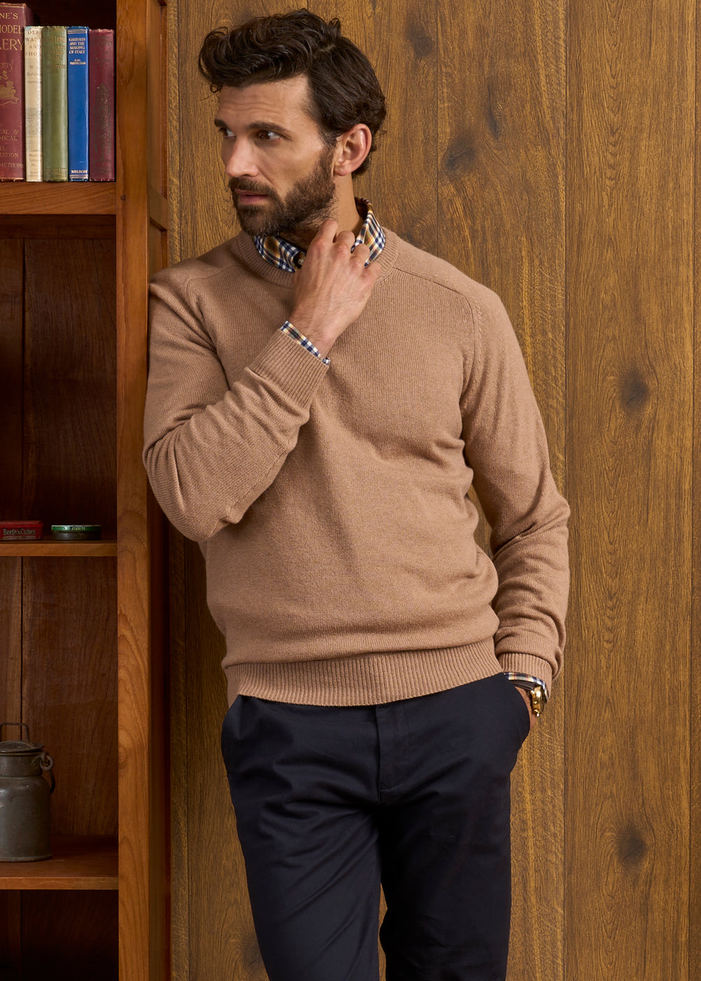 Man in Lenzie Lambswool Crew Neck Jumper in Camel stands by bookshelf, looking left with hand on neck.