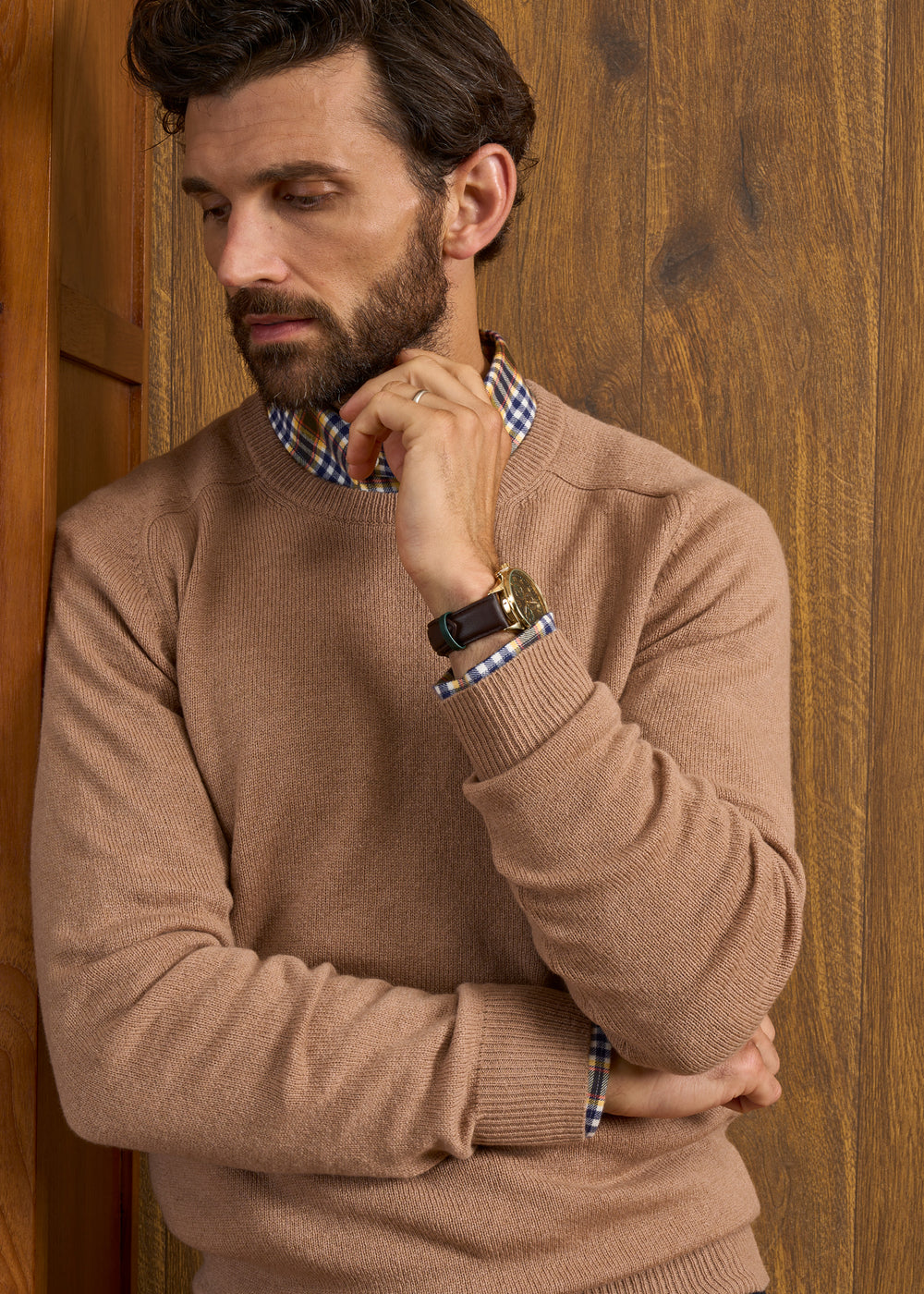 Man in Lenzie Lambswool Crew Neck Jumper in camel stands by wood wall, looking down with hand near his face.