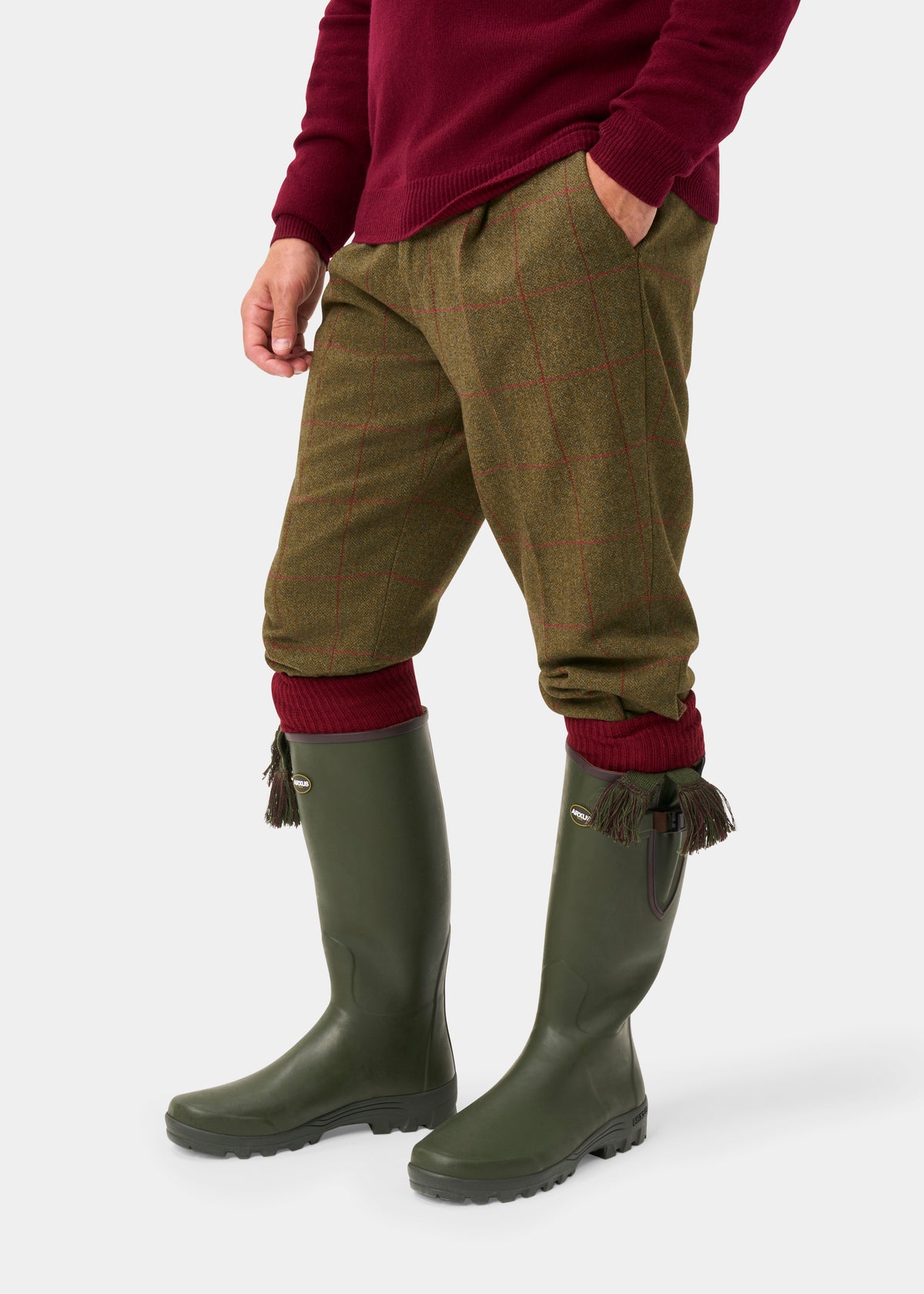 Dressed in sage tweed breeks and green boots, he stands against a white background in a burgundy sweater.