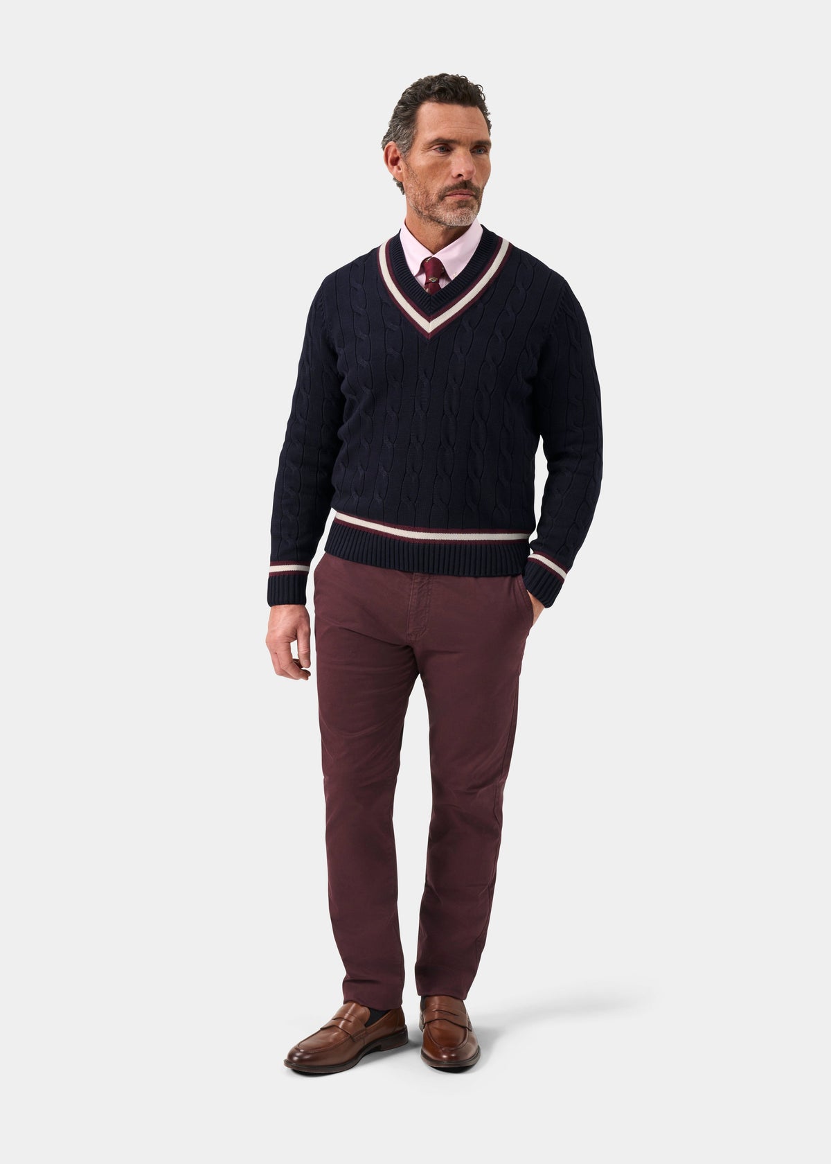 A man wears a Sandridge Cotton Cable Knit V-Neck Cricket Jumper In Dark Navy and Blackcurrant over a pink shirt and trousers.
