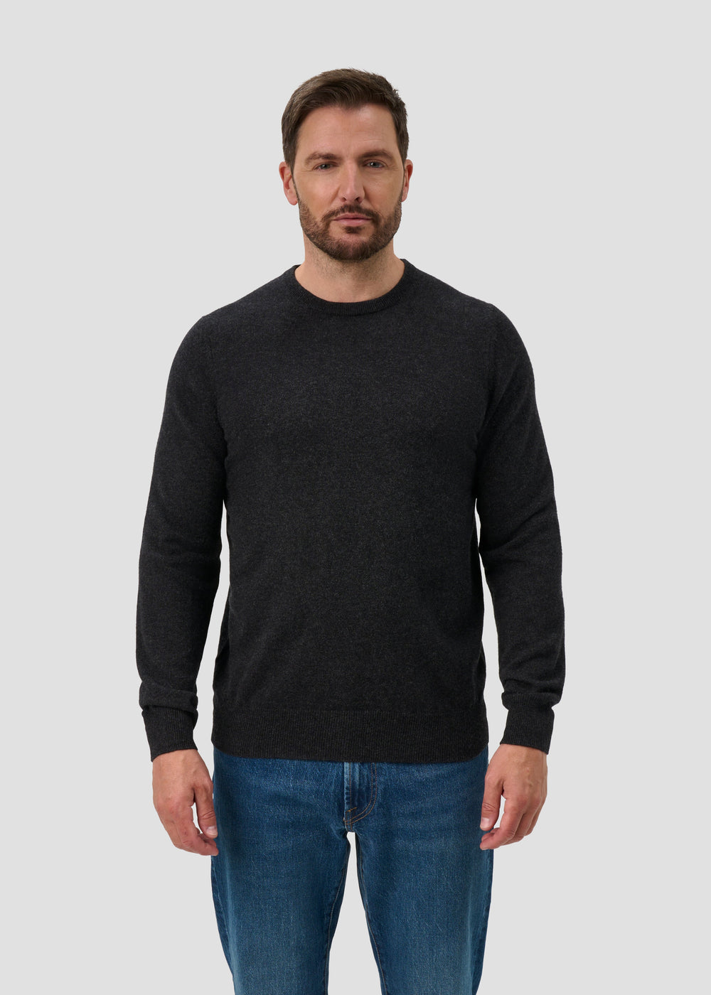 Man with short brown hair and beard wears Brisbane Geelong Wool Crew Neck Jumper in charcoal and blue jeans.