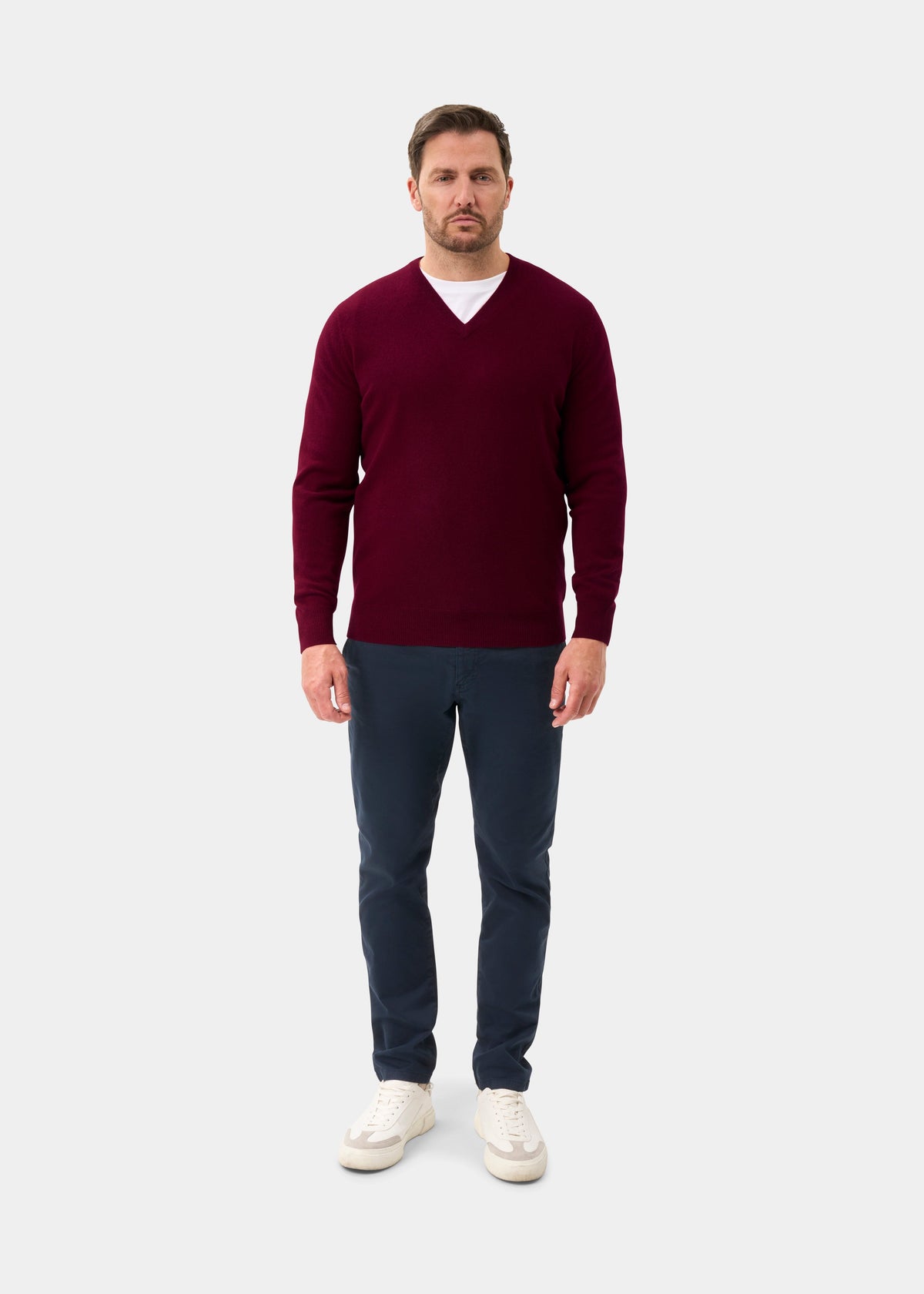 Man in Albury Geelong Wool V-Neck Jumper in Claret, white shirt, navy pants, and white sneakers on light background.