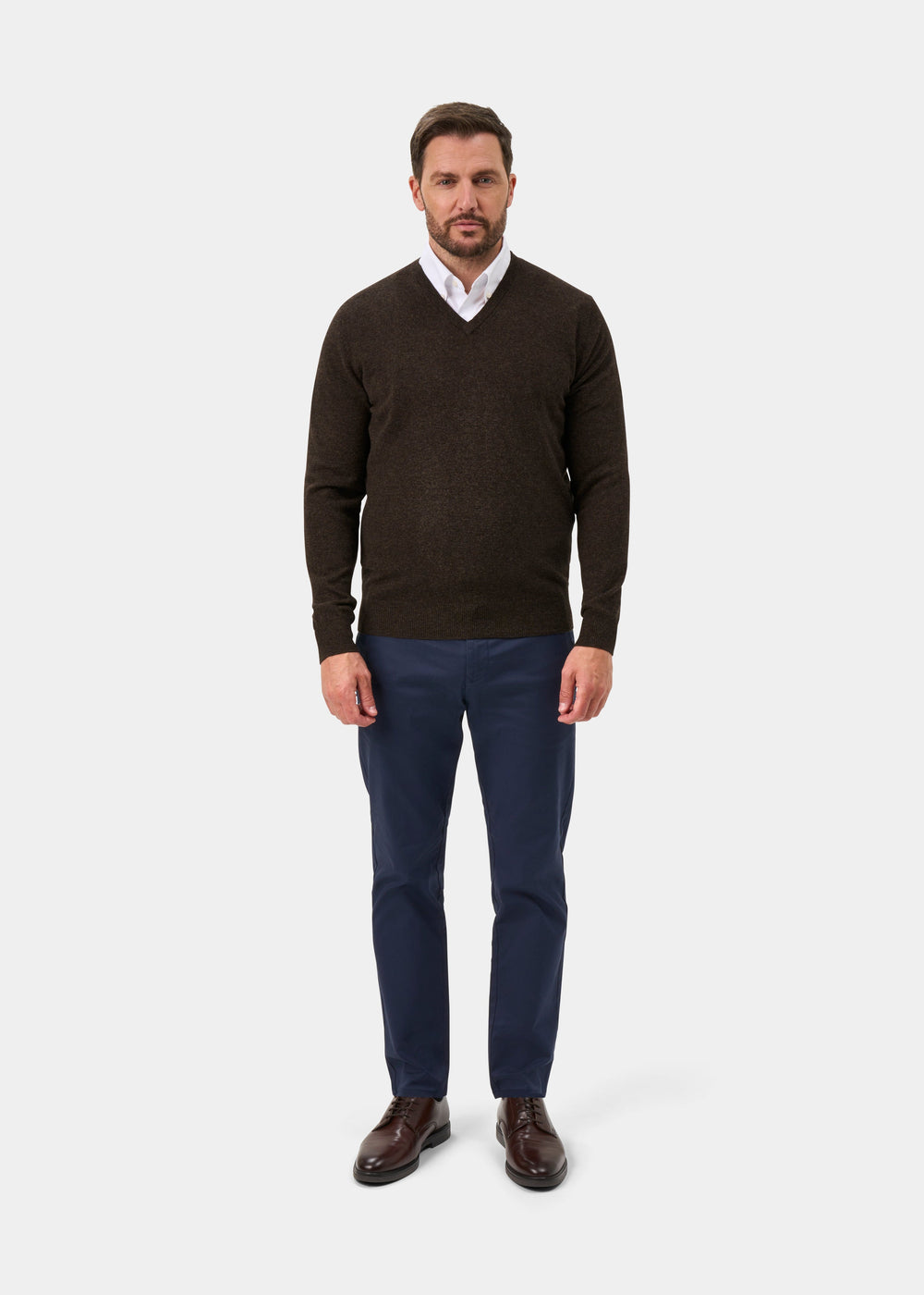 Man wears Albury Geelong Wool V-Neck Jumper in Cocoa with white shirt, blue pants, brown shoes; faces plain background.