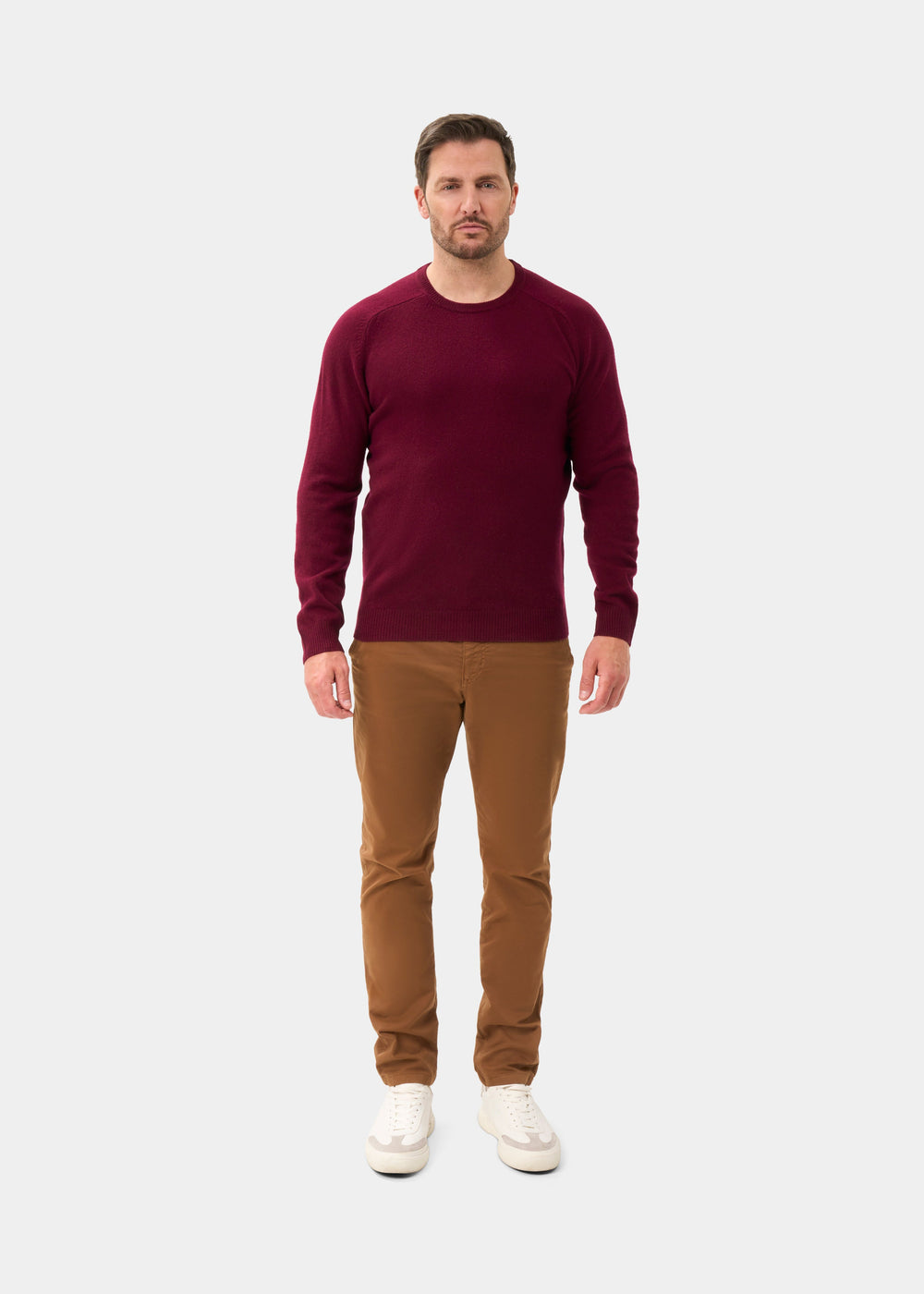 Man in Lenzie Lambswool Crew Neck Jumper in Bordeaux, brown pants, and white shoes on a plain white background.