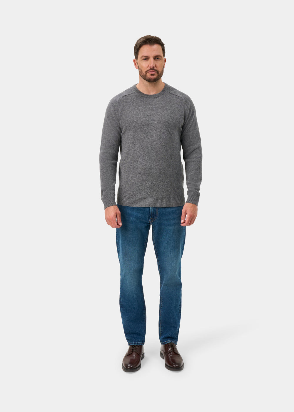 Man wears Lenzie Lambswool Crew Neck Jumper in Grey Mix, blue jeans, and brown shoes, facing forward on white background.