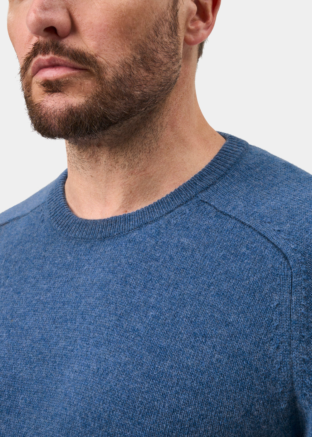 A bearded man wears the Lenzie Lambswool Crew Neck Jumper in Jeans, shown from shoulders up on a white background.
