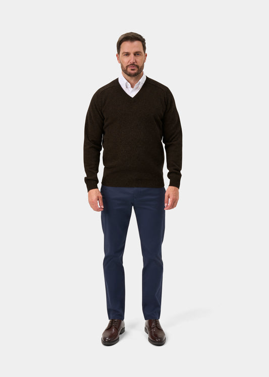 Man in Kilsyth Lambswool Vee Neck Jumper in Cocoa, white shirt, blue pants, brown shoes stands against a white background.