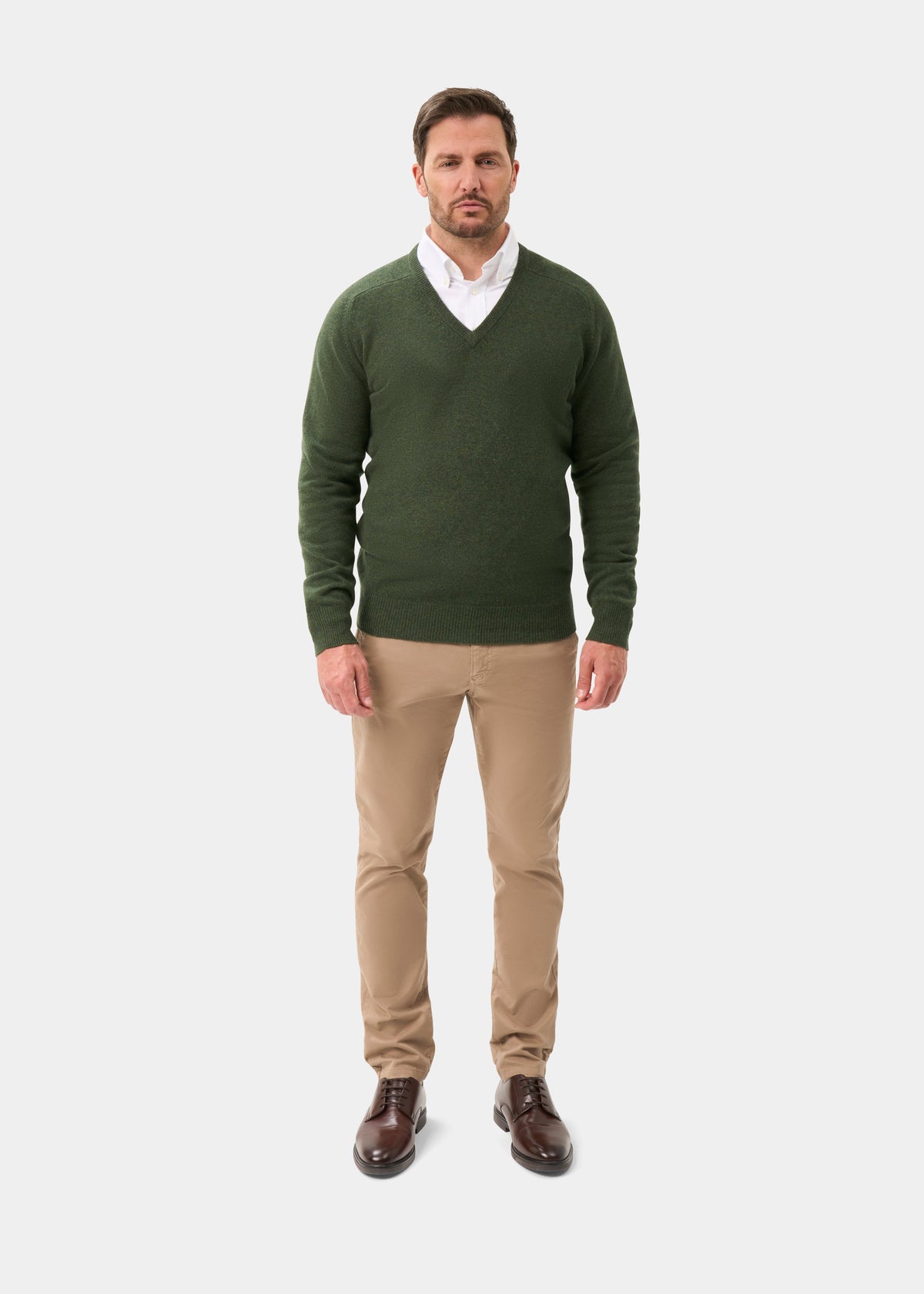 Man in Hampshire Lambswool V-Neck Jumper in Rosemary, white shirt, beige pants, brown shoes on white background.