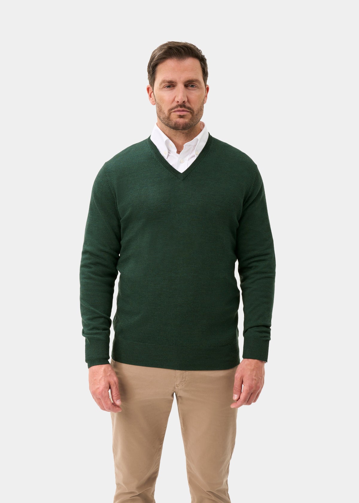 A man wearing a dark green v-neck knit sweater over a white collared shirt and beige trousers, standing against a light gray studio background.