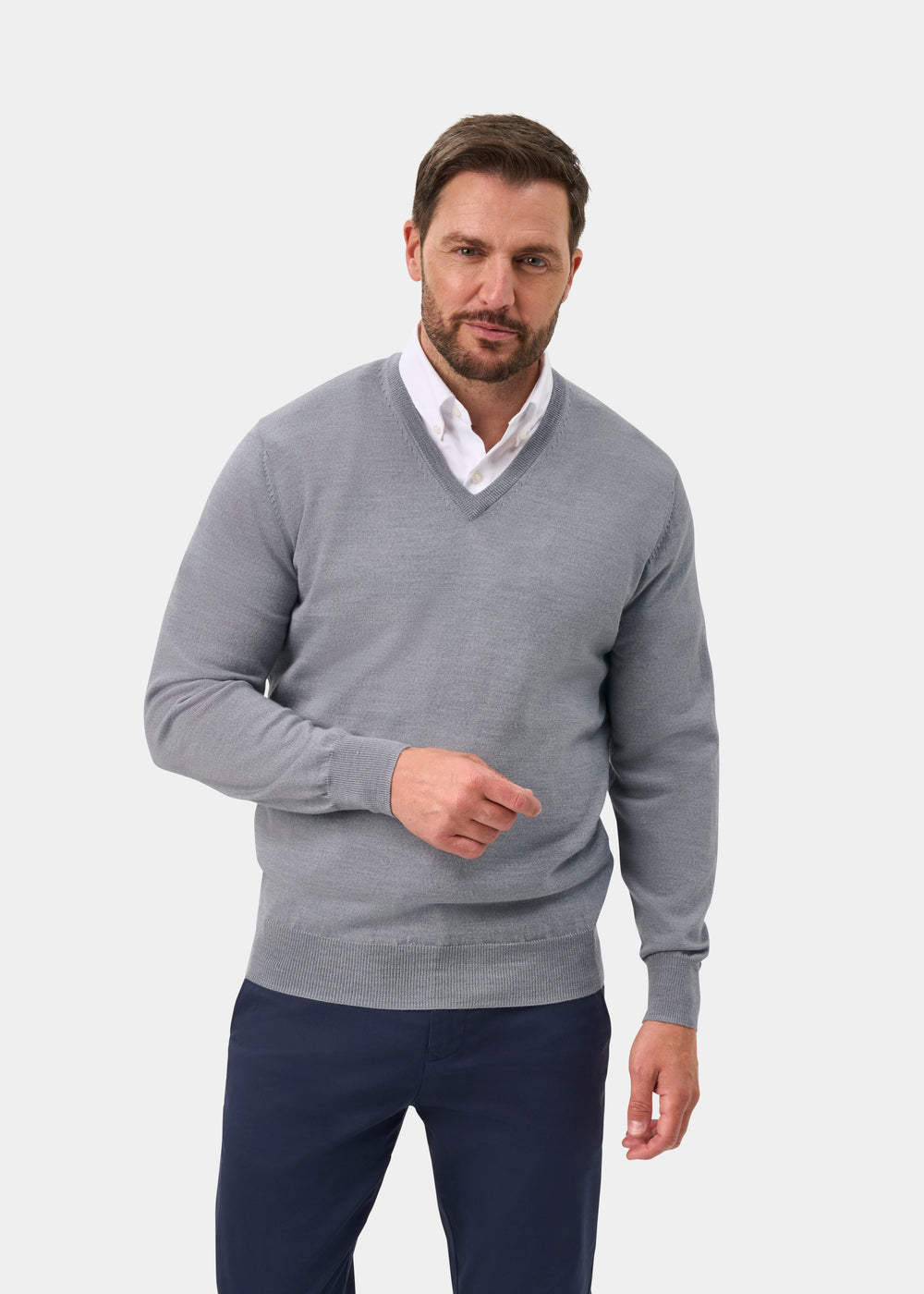 A man wearing a light gray v-neck knit sweater over a white collared shirt and navy trousers, standing against a plain light gray studio background.