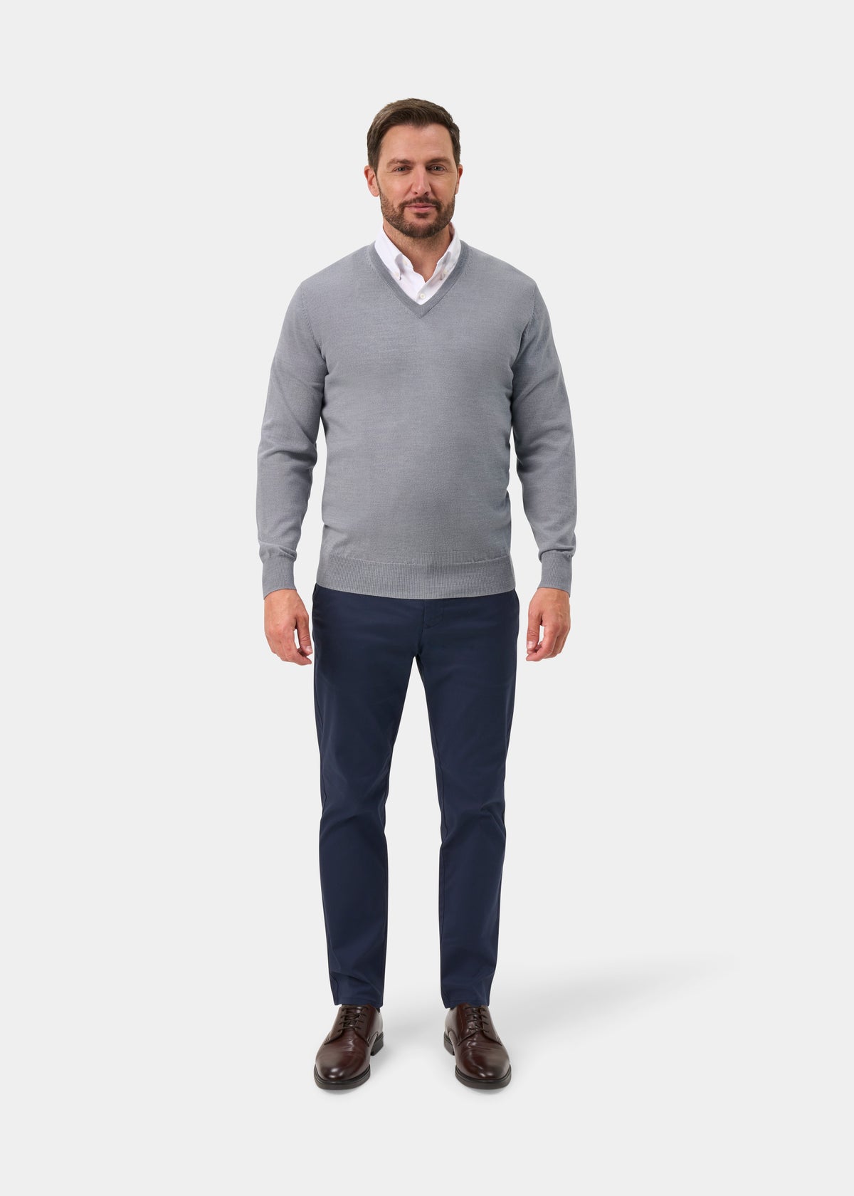 A man wearing a light gray v-neck sweater over a white collared shirt, paired with navy blue trousers and dark brown dress shoes, standing against a light gray studio background.