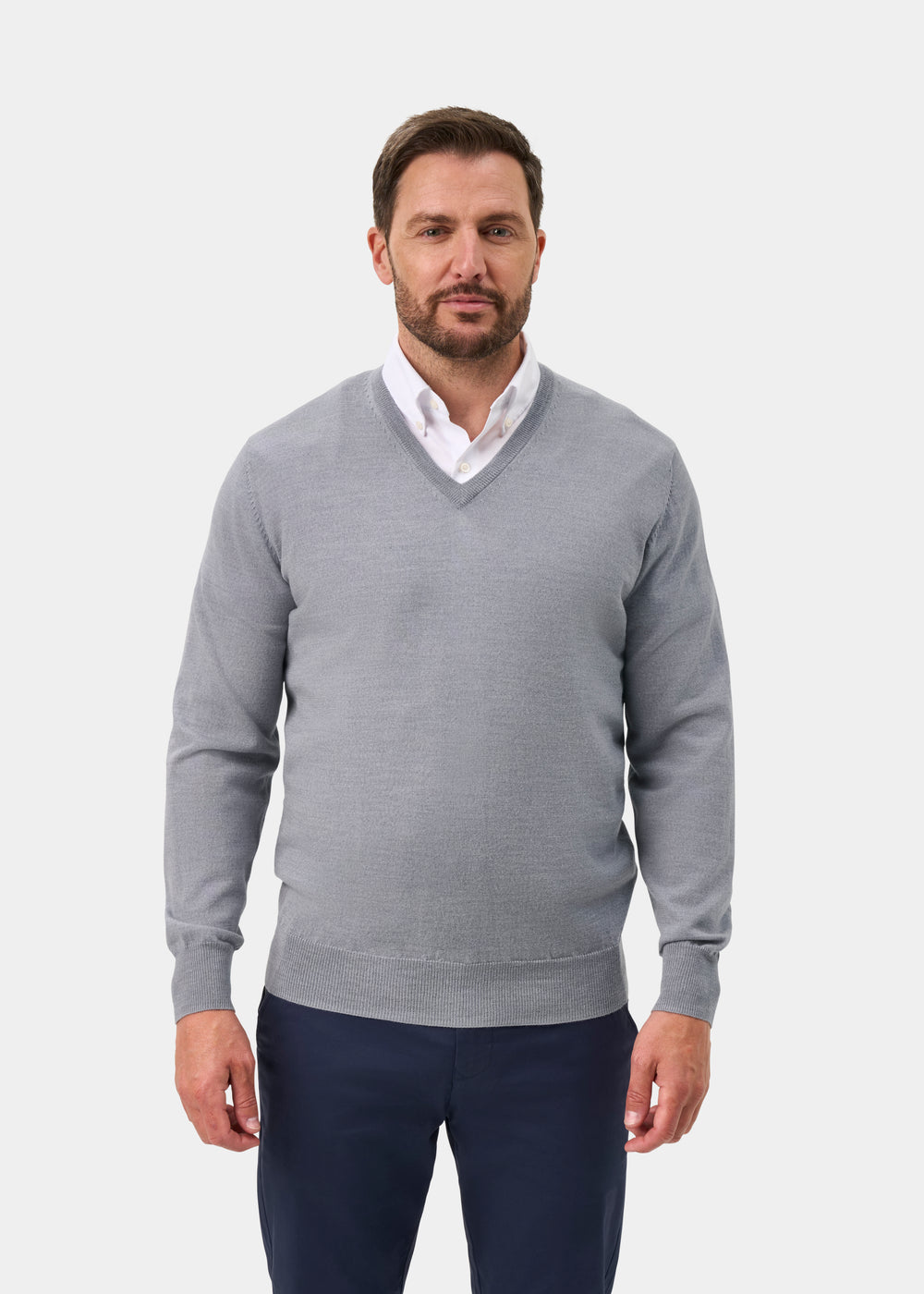A man wearing a light gray v-neck sweater over a white collared shirt and navy trousers, standing against a plain light gray studio background.