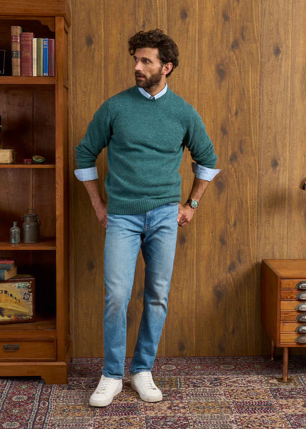 A man in a Kinnadie Supersoft Shetland Wool Jumper, blue shirt, jeans, and white sneakers stands by wooden furniture.