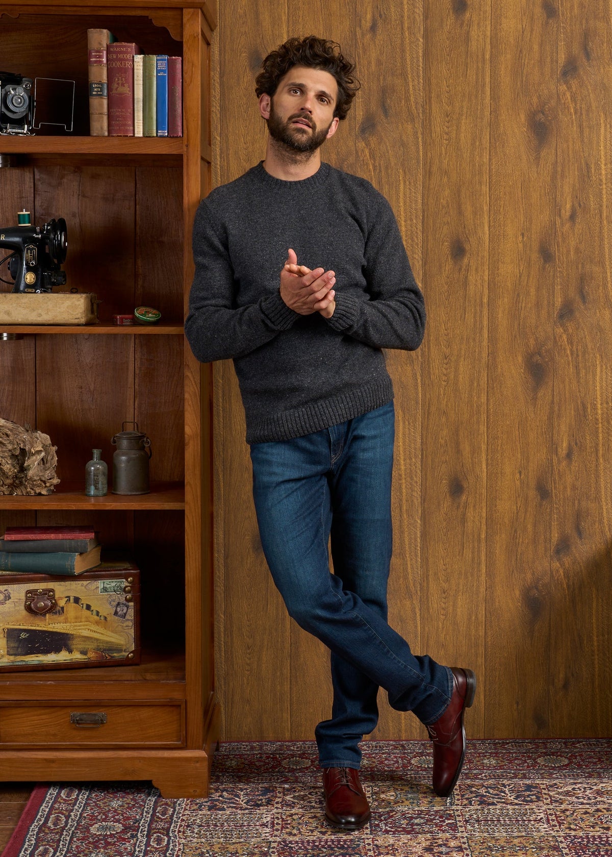 Man in Kinnadie Oxford Pine Shetland Wool Jumper, jeans by wooden shelf, hands clasped on patterned rug, wood wall.
