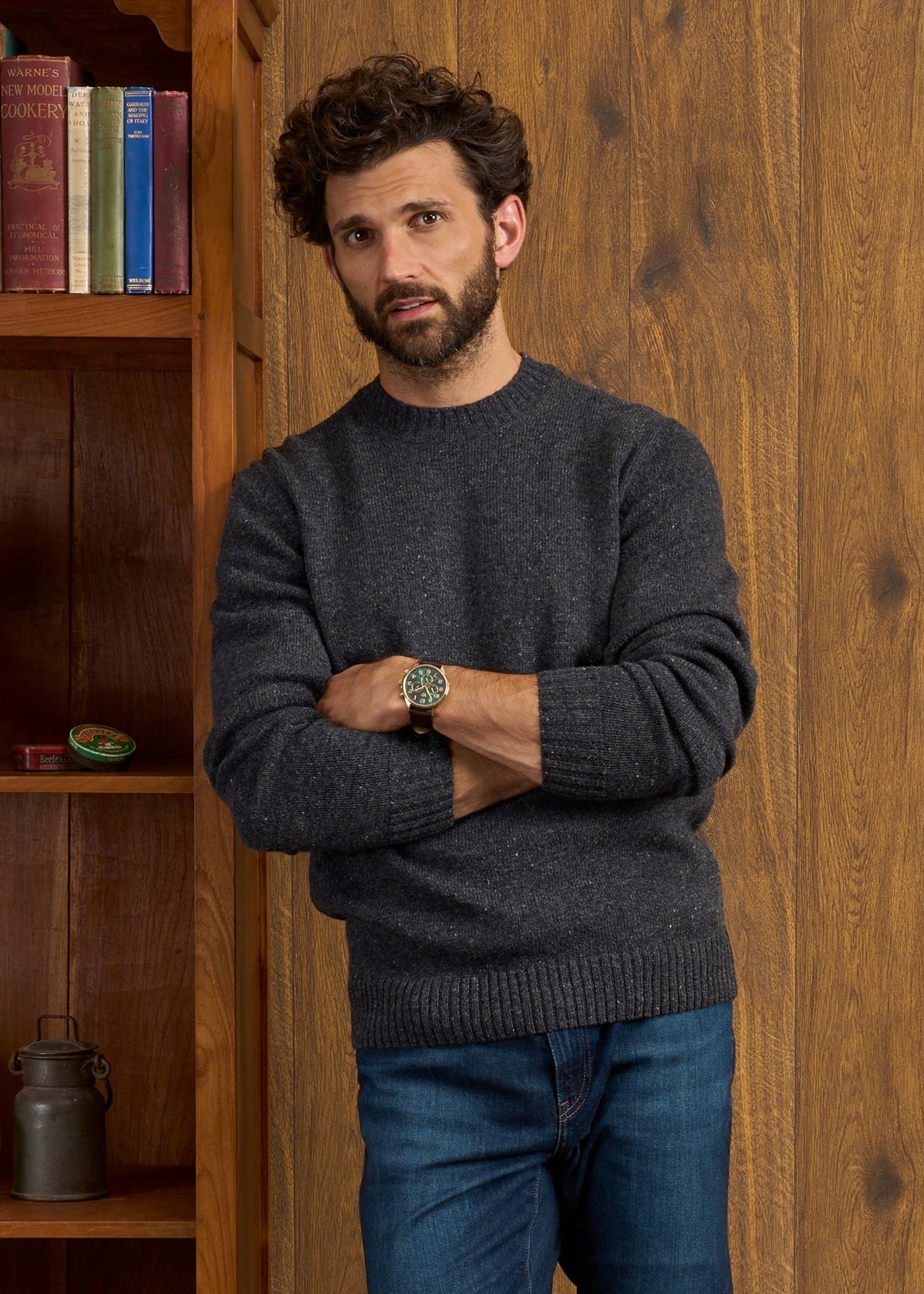 Man with curly hair and beard wears the Kinnadie Supersoft Shetland Wool Jumper in Oxford Pine, arms crossed by bookshelf.