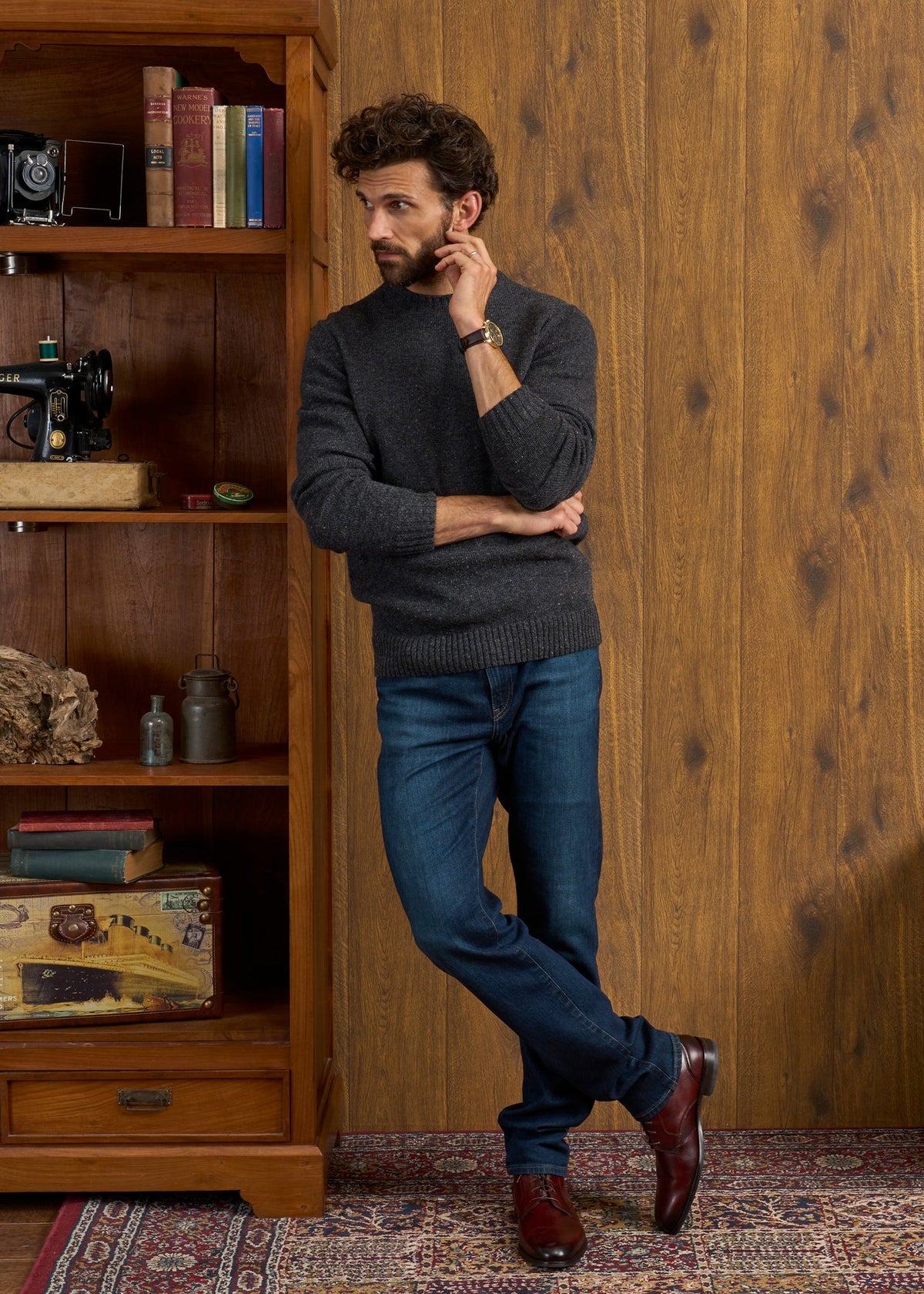 Man in a Kinnadie Shetland Wool Jumper in Oxford Pine and jeans leans on a bookshelf, standing on a patterned rug.