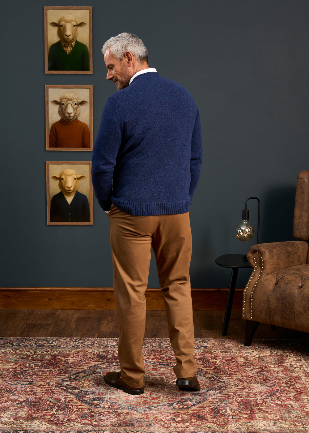 A man in a Kinnadie Shetland Wool Crew Neck Jumper in Indigo and tan pants stands on a rug, facing sheep portraits.