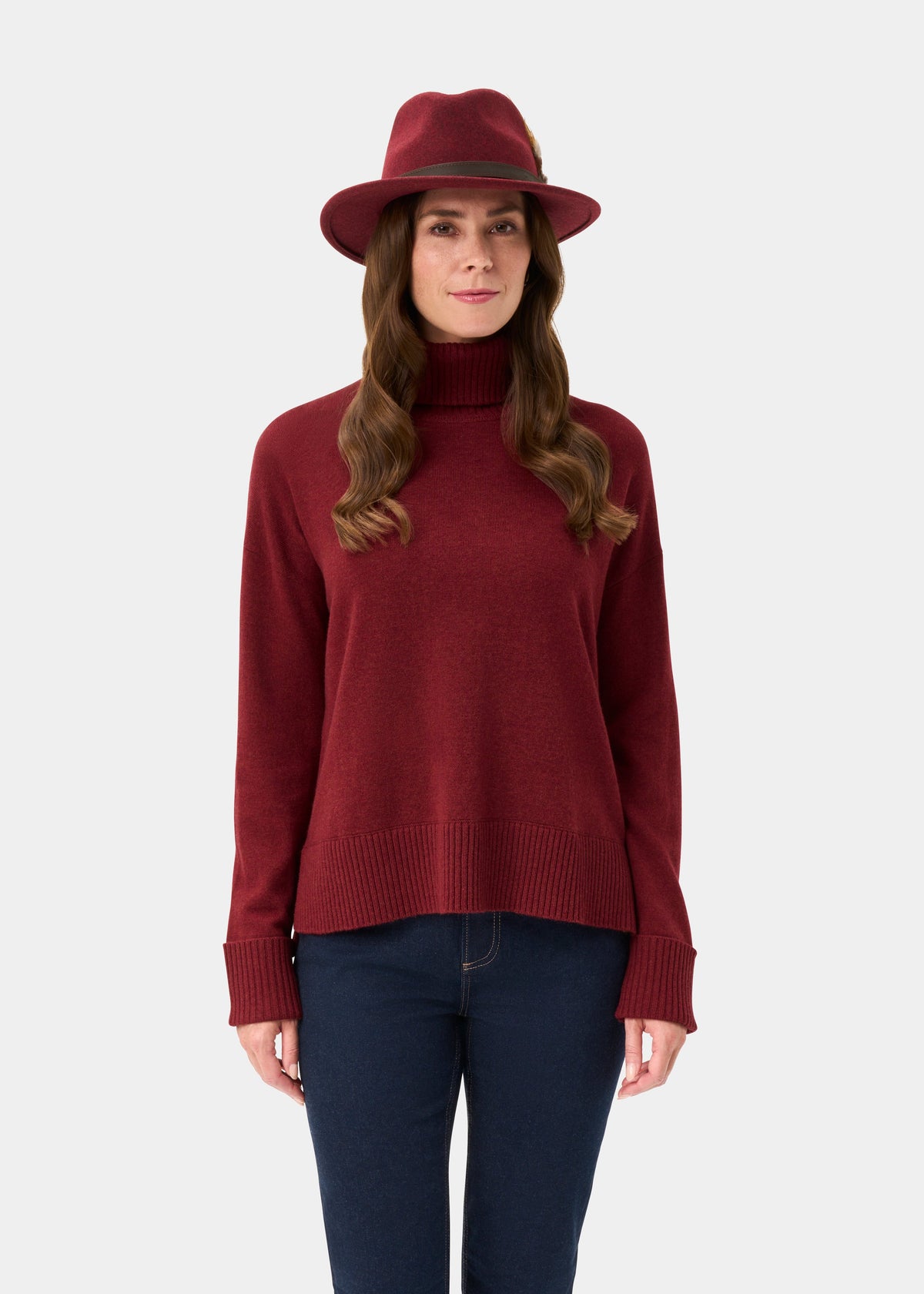 Woman in Alexa Ladies Wool and Cashmere Roll Neck in Berry, red hat, and blue jeans stands against a plain background.