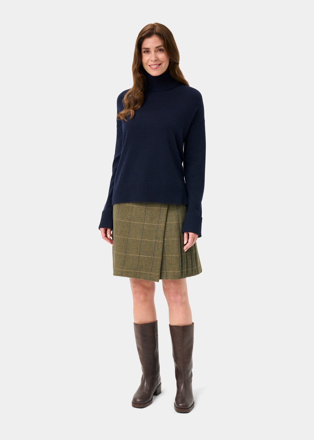 Woman in an Alexa Wool & Cashmere Roll Neck Jumper in dark navy, green plaid skirt, and brown boots, smiling.