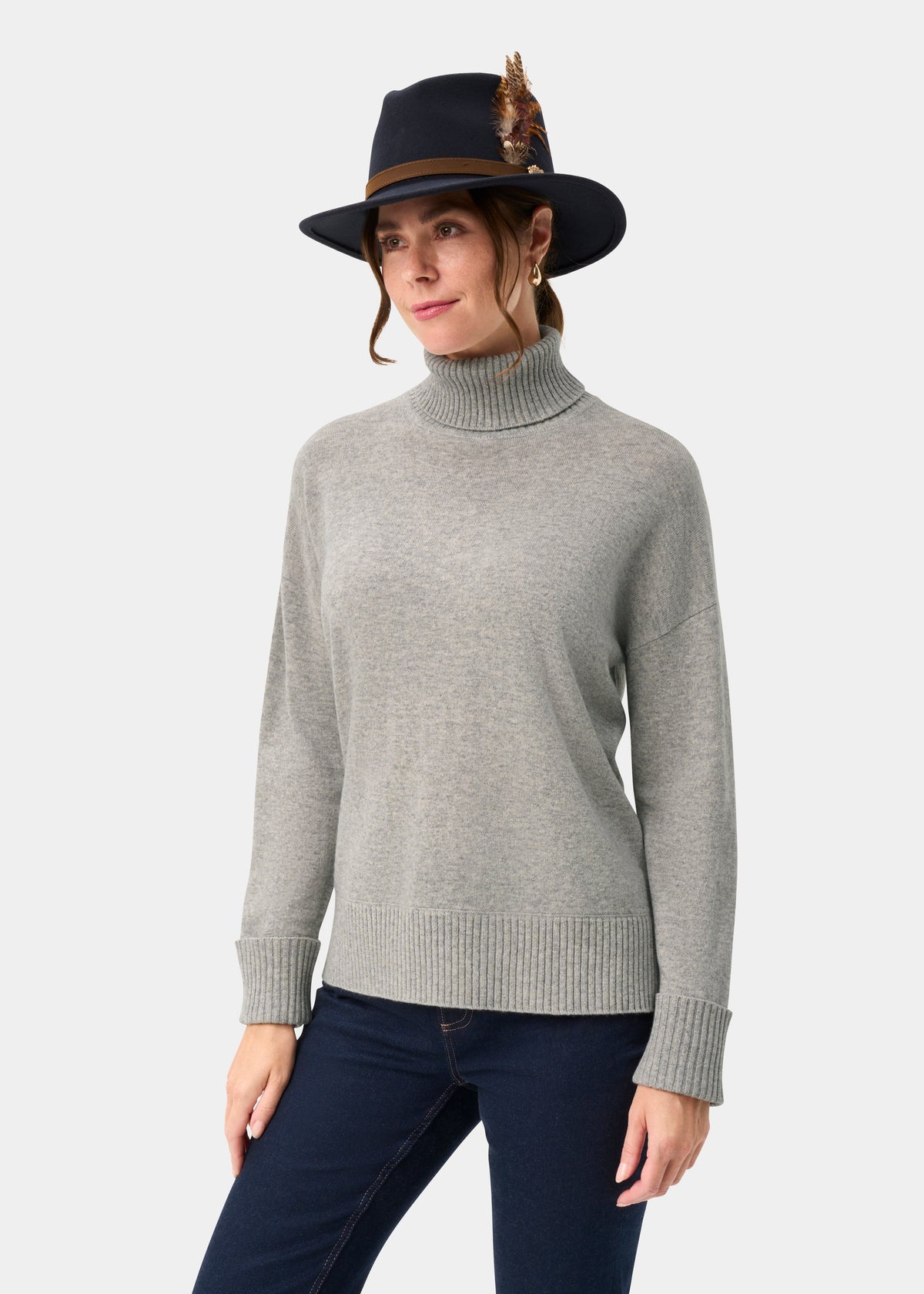 Woman wears Alexa Wool & Cashmere Roll Neck Jumper in Husky, dark jeans, and a black feathered hat against plain backdrop.