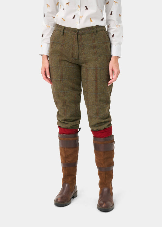 A model is shown from the waist down wearing olive green plaid tweed trousers, red knit socks, and brown riding boots, paired with a white patterned shirt, against a light gray background.