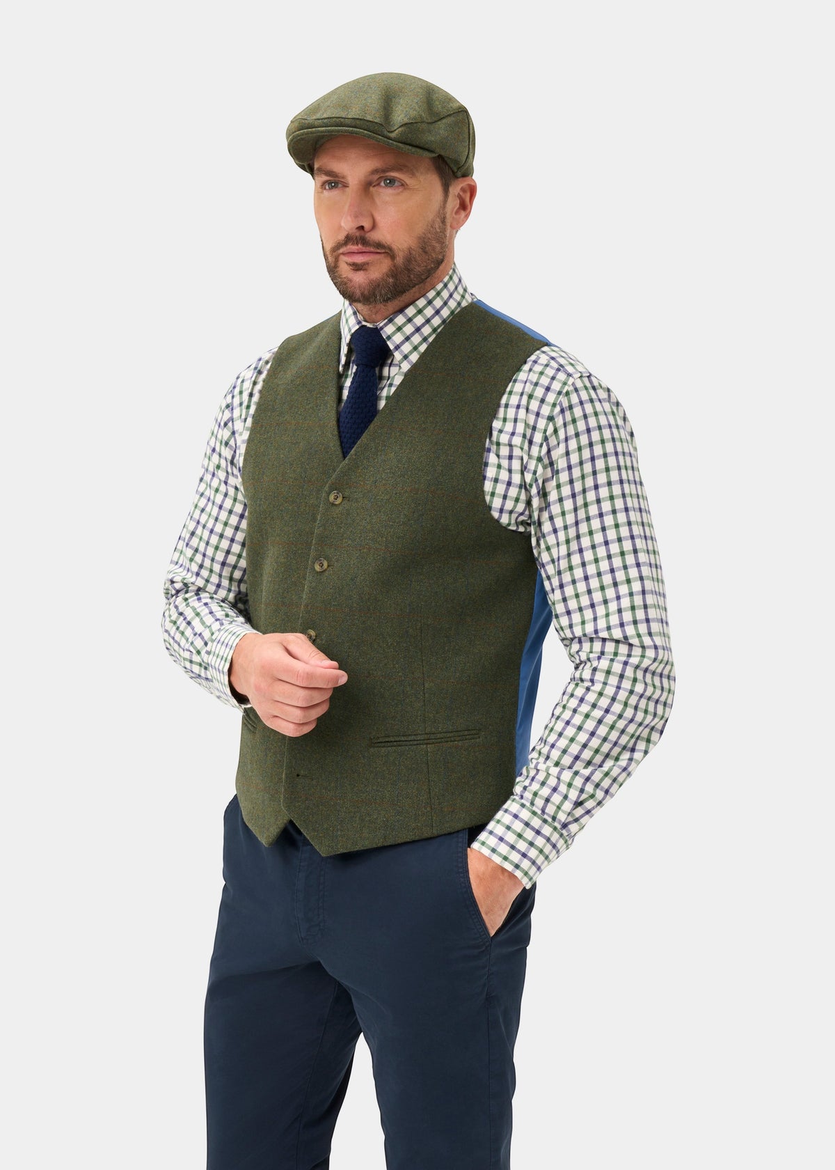 Man wears Combrook Men’s Mariner Tweed Waistcoat, flat cap, plaid shirt, navy tie and pants, hand in pocket.