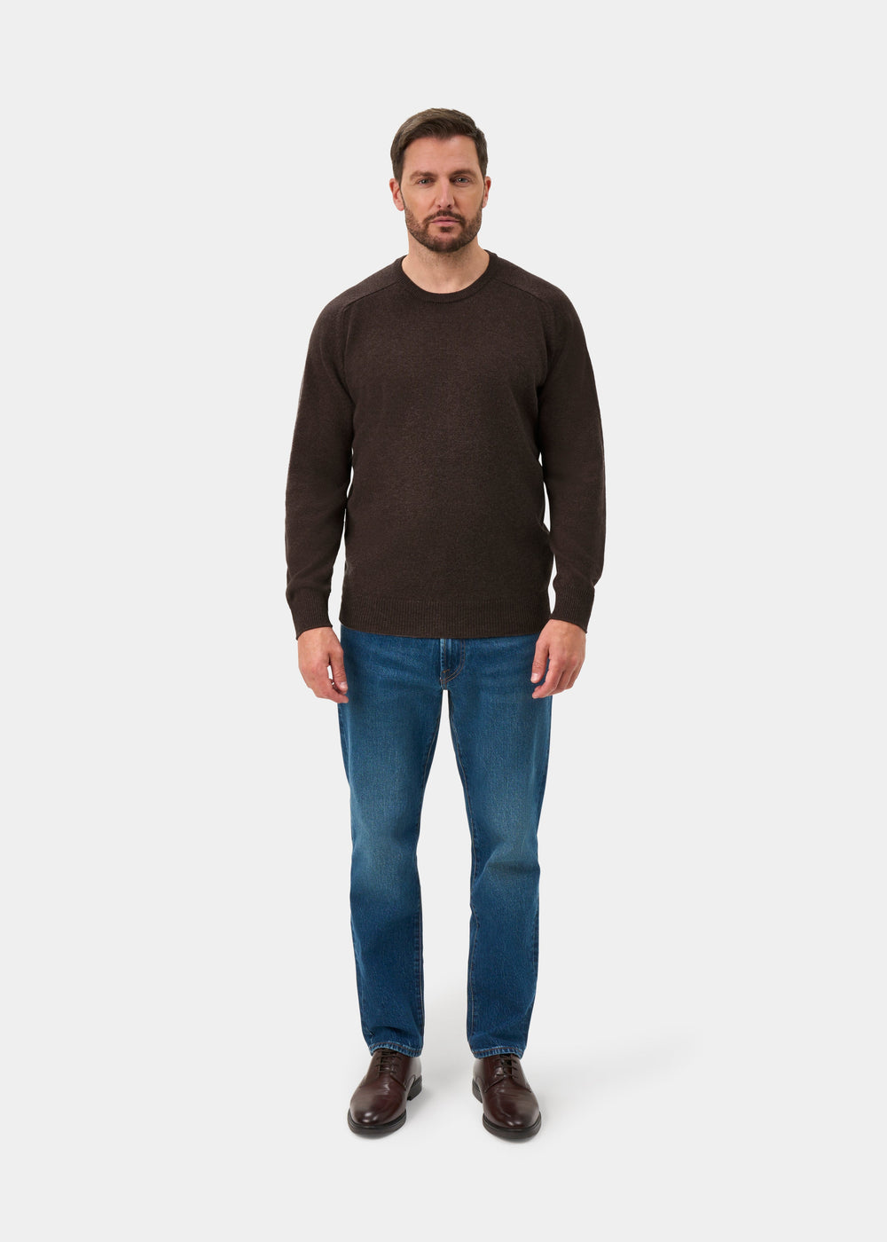 Man wearing Dorset Lambswool Crew Neck Jumper in mocha, blue jeans, and brown shoes on a plain white background.