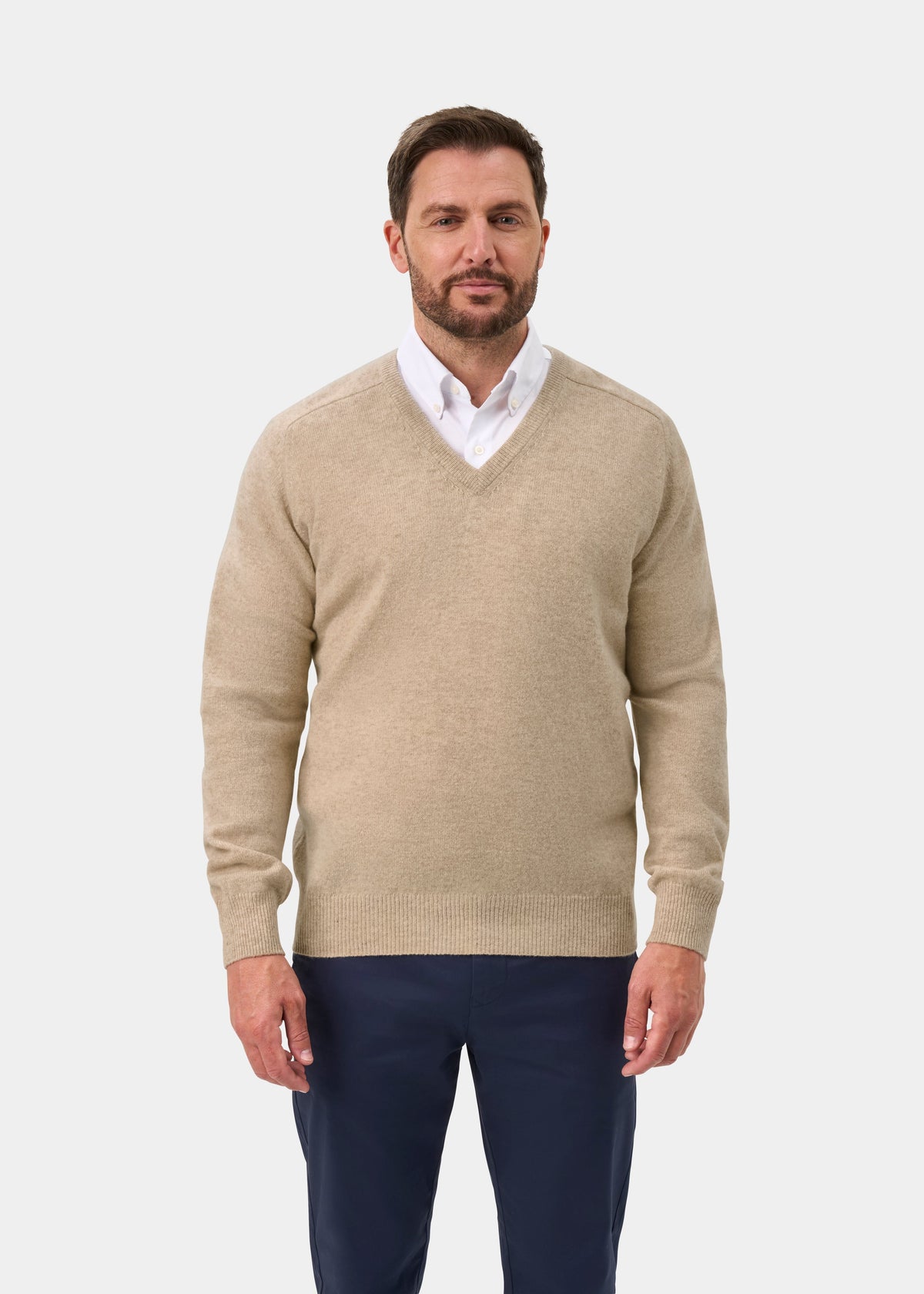 Man wears Hampshire Lambswool V-Neck Jumper in Linen over a white shirt and navy pants, against white background.