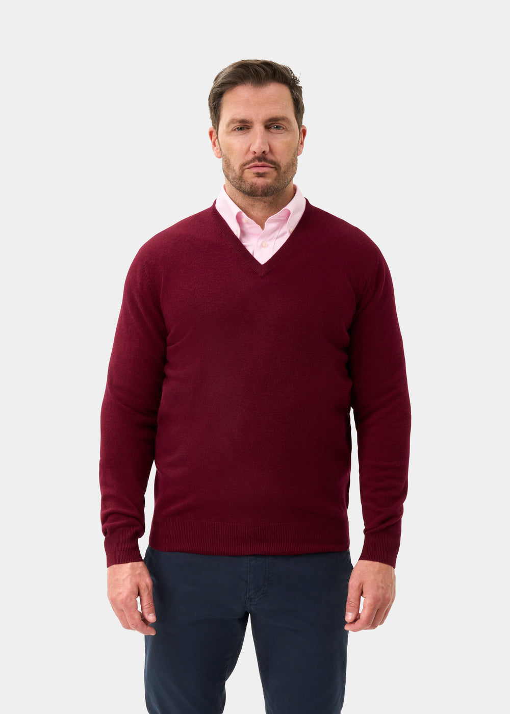 Man wearing Kilsyth Lambswool V-Neck Jumper in Garnet over a pink shirt and navy pants, on a white background.