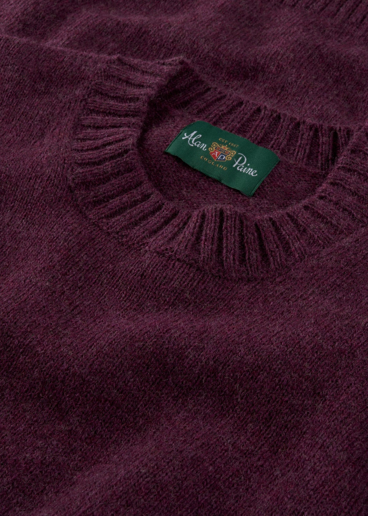 A close-up of the Kinnadie Shetland Wool Crew Neck Jumper in Elderberry shows its ribbed collar and green label.