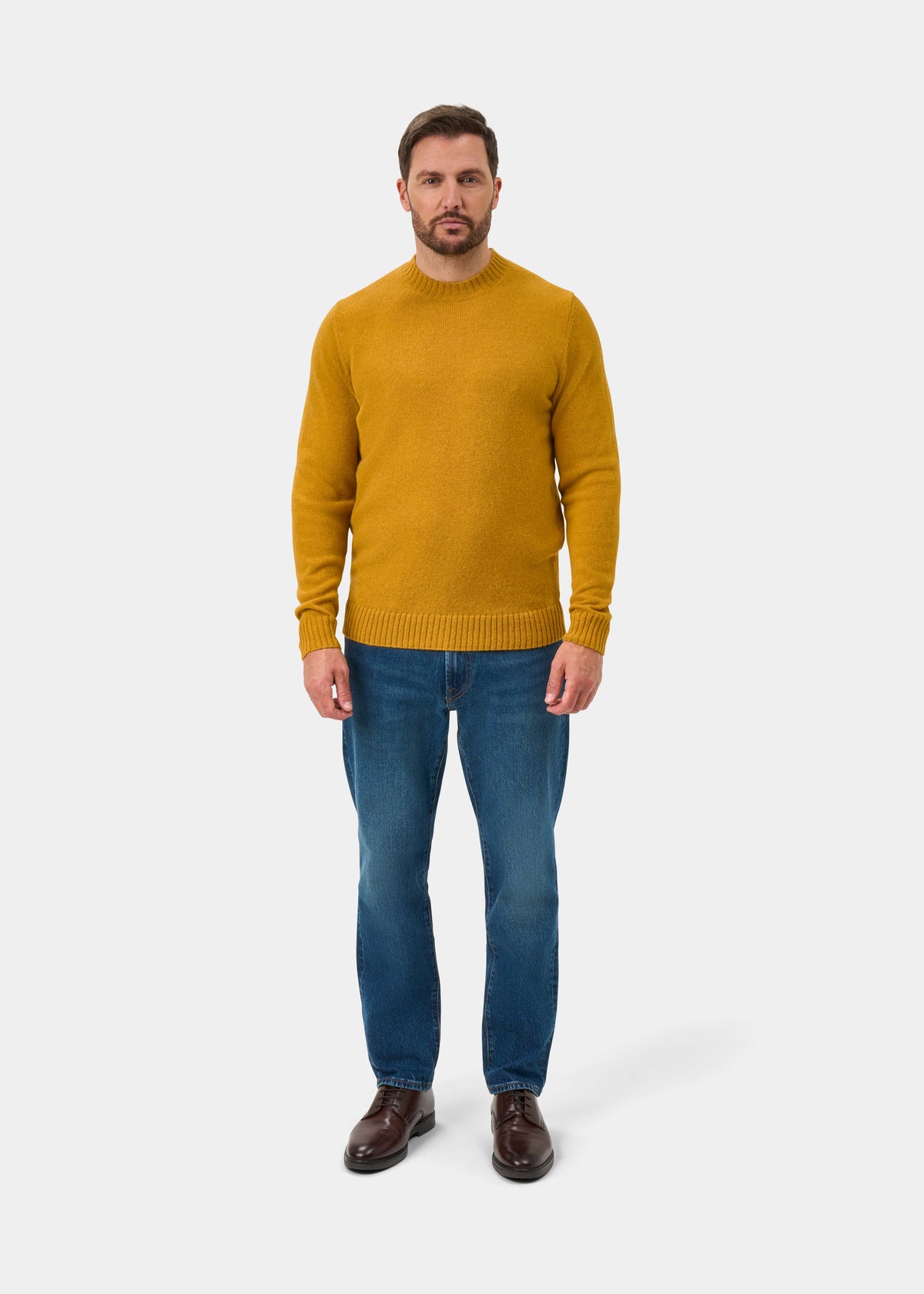 Man in a Kinnadie Old Gold Supersoft Shetland Wool Jumper, blue jeans, and brown shoes stands against a white backdrop.