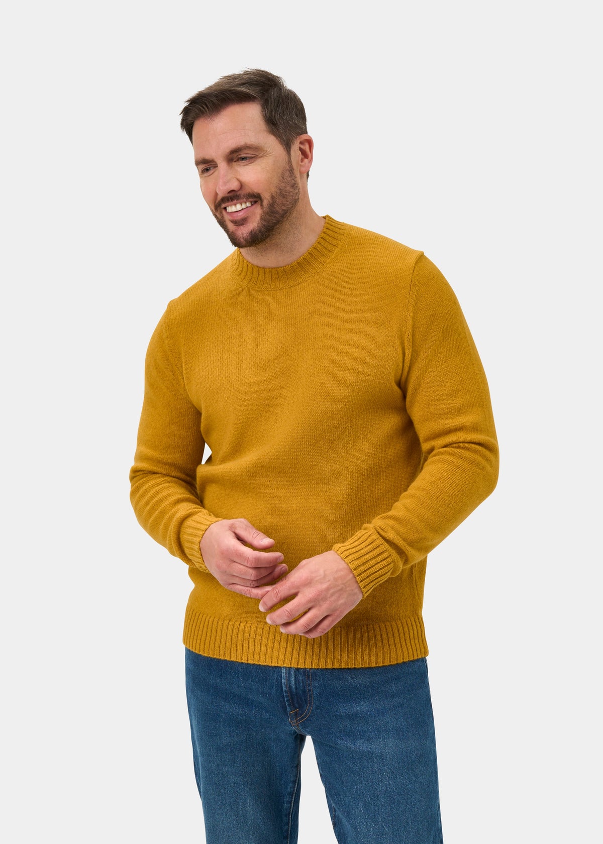 Smiling man in Kinnadie Supersoft Shetland Wool Jumper in Old Gold, blue jeans, hands raised on white background.